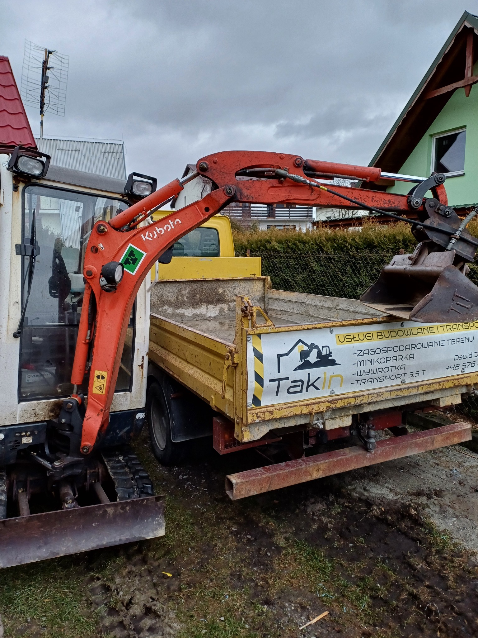 Mini koparka Kubota z pomarańczowym ramieniem i łyżką, obok żółta wywrotka z reklamą firmy Takin, na błotnistym podłożu w pochmurny dzień.