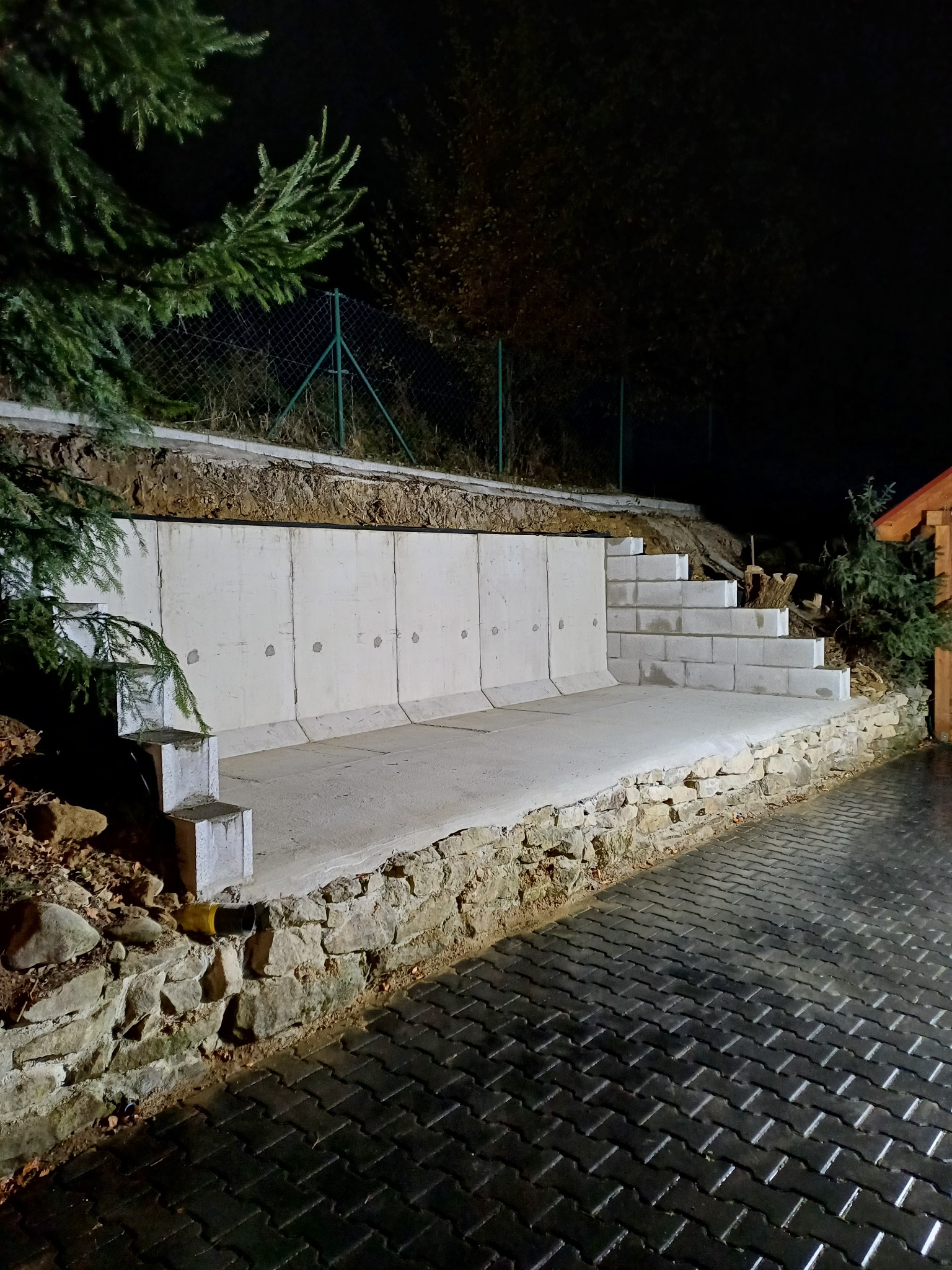 Mur oporowy z betonowych elementów z niskimi schodami, podtrzymujący skarpę, wykończony kamienną podmurówką, widok na mokrą kostkę brukową i ogrodzenie z siatki w tle, nocne oświetlenie.