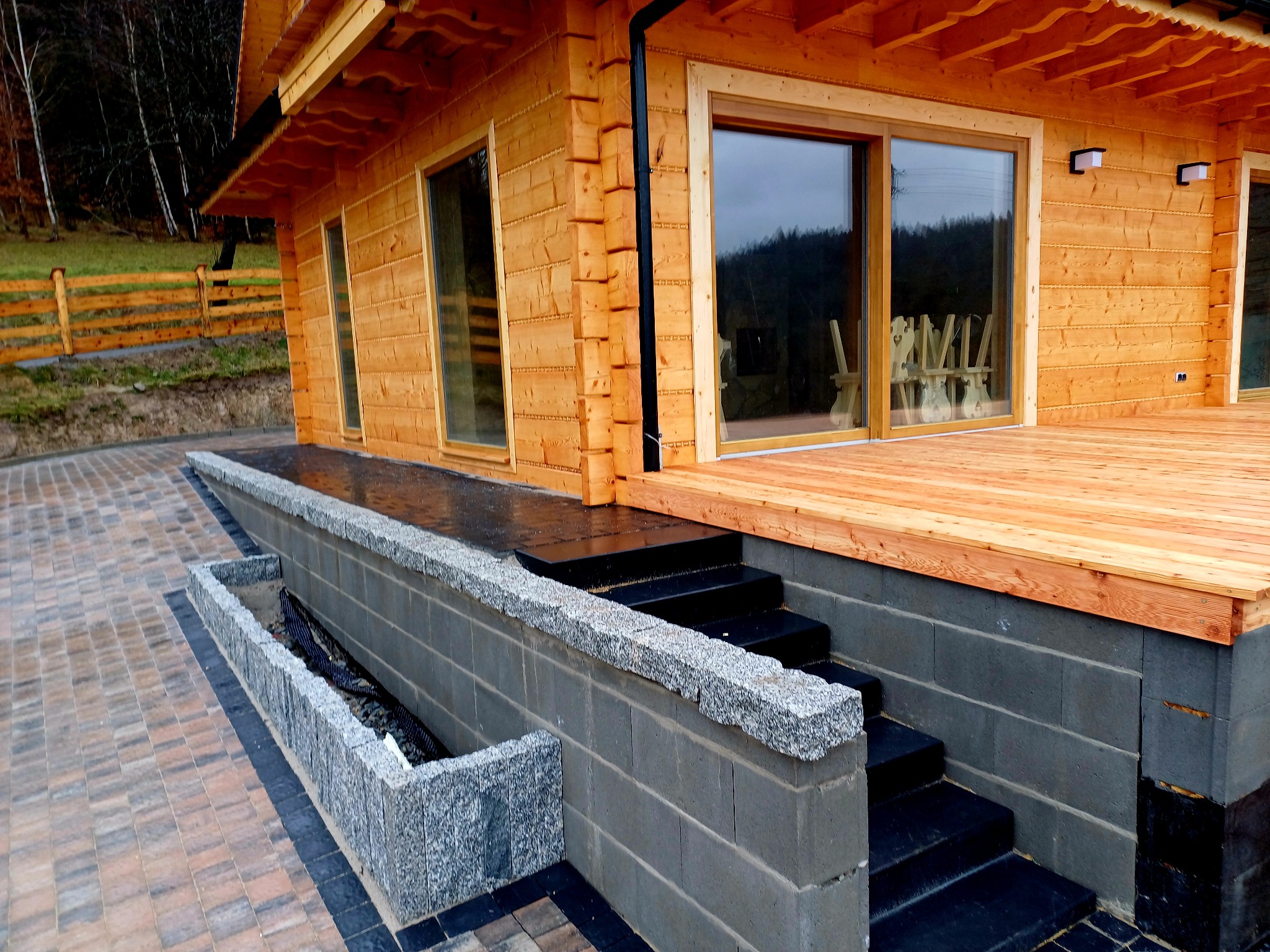 Nowoczesny dom z bali, z tarasem wyłożonym drewnem i schodami z czarnego kamienia, otoczony kostką brukową i granitowym obrzeżem, w tle zielony krajobraz z drewnianym płotem.