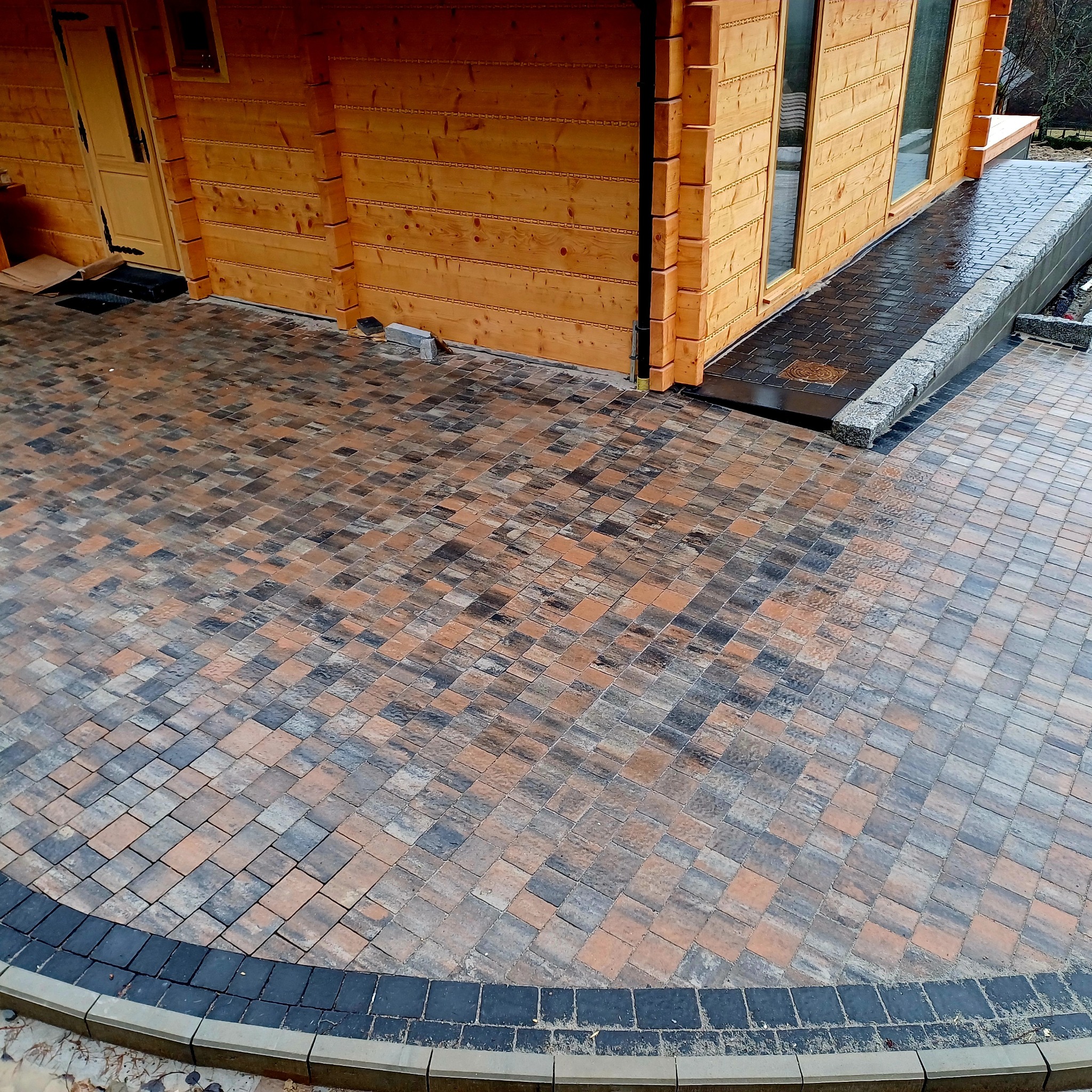 Ułożona kostka brukowa w odcieniach brązu i szarości przed drewnianym domem, z obrzeżem z ciemnej kostki i szarego betonu.