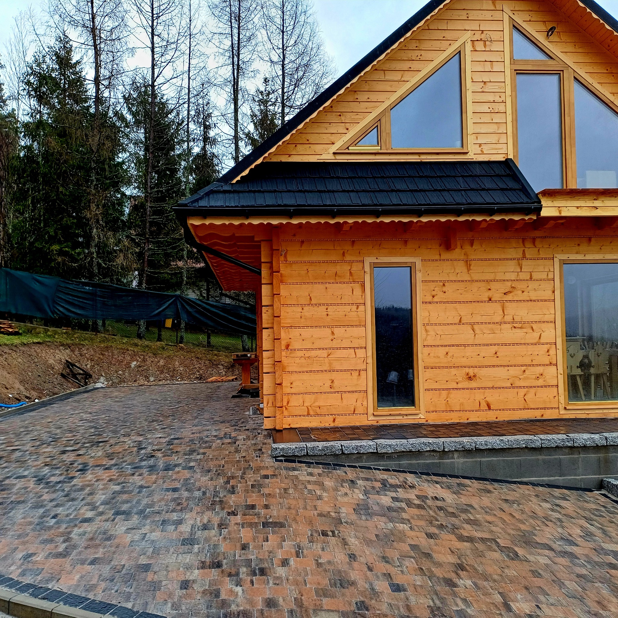 Nowo ułożona, wielobarwna kostka brukowa prowadząca do drewnianego domu z oknami o nietypowym kształcie, widoczny fragment posesji z drzewami w tle.