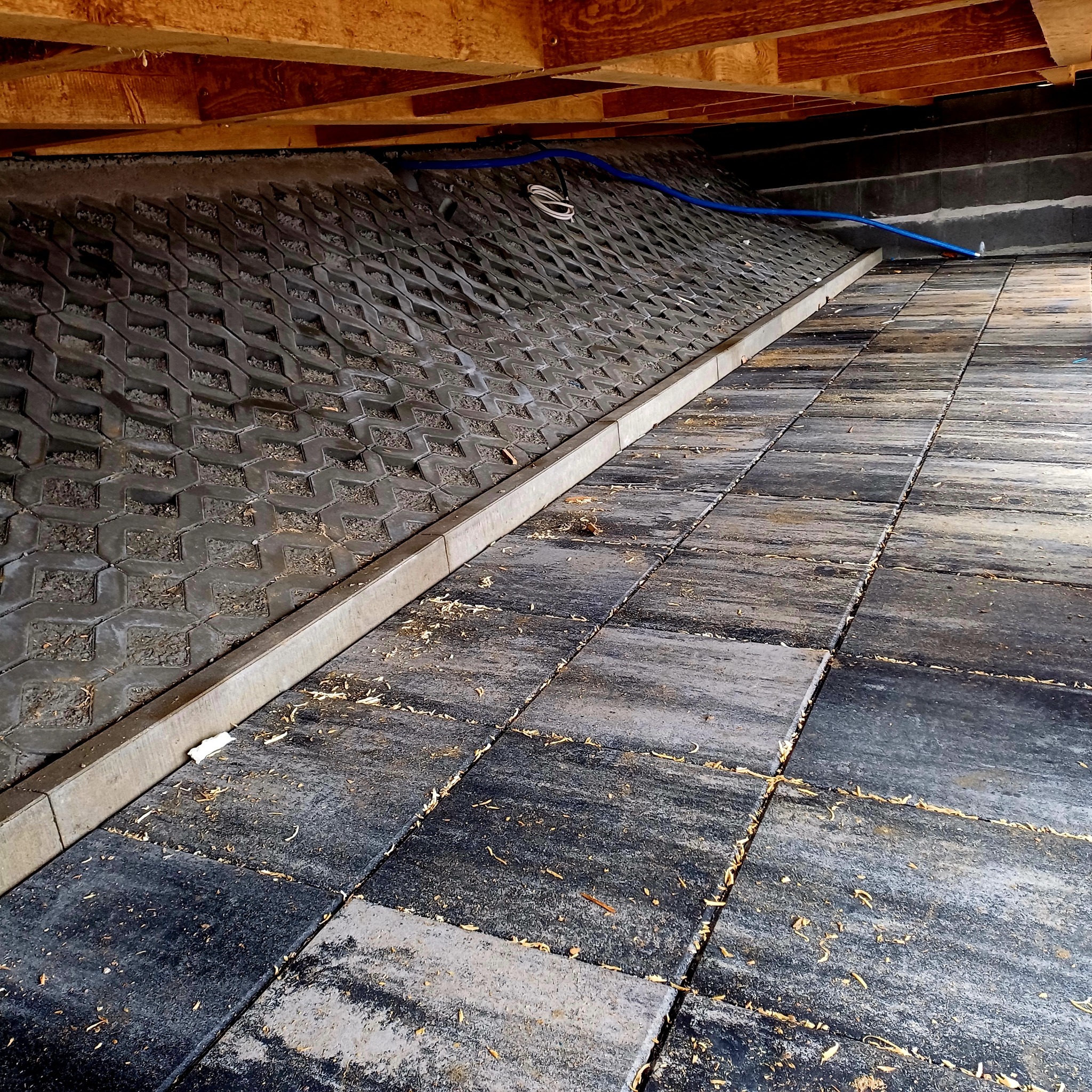 Ułożona nawierzchnia z kostki brukowej w odcieniach szarości, z widocznym fragmentem ażurowej kostki na skarpie, ograniczonej betonowym obrzeżem i z wystającymi przewodami instalacyjnymi...