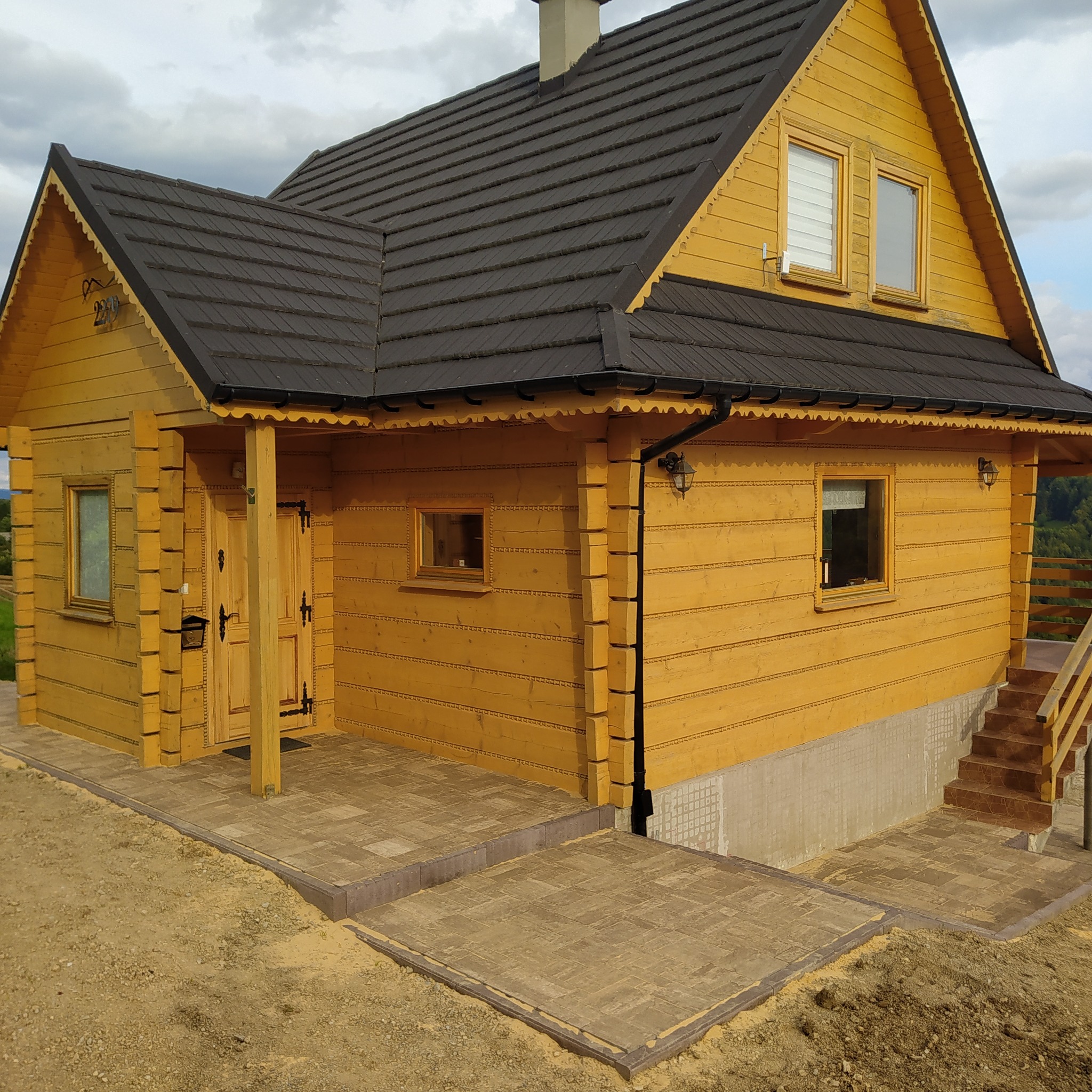 Żółty dom z bali z ciemną dachówką, widoczne schody i fragment chodnika z kostki brukowej przed wejściem, otoczenie w stanie surowym.