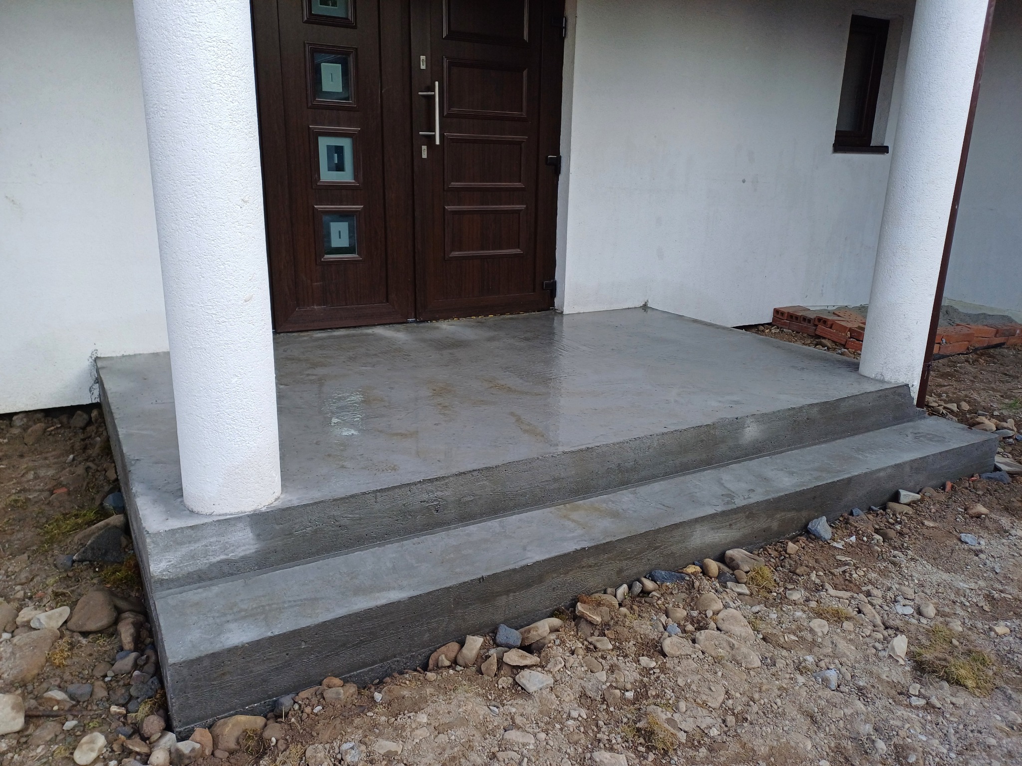 Nowy, betonowy podest wejściowy z dwoma stopniami, wykończony przy białych kolumnach i brązowych drzwiach wejściowych. Otoczenie w trakcie zagospodarowania.