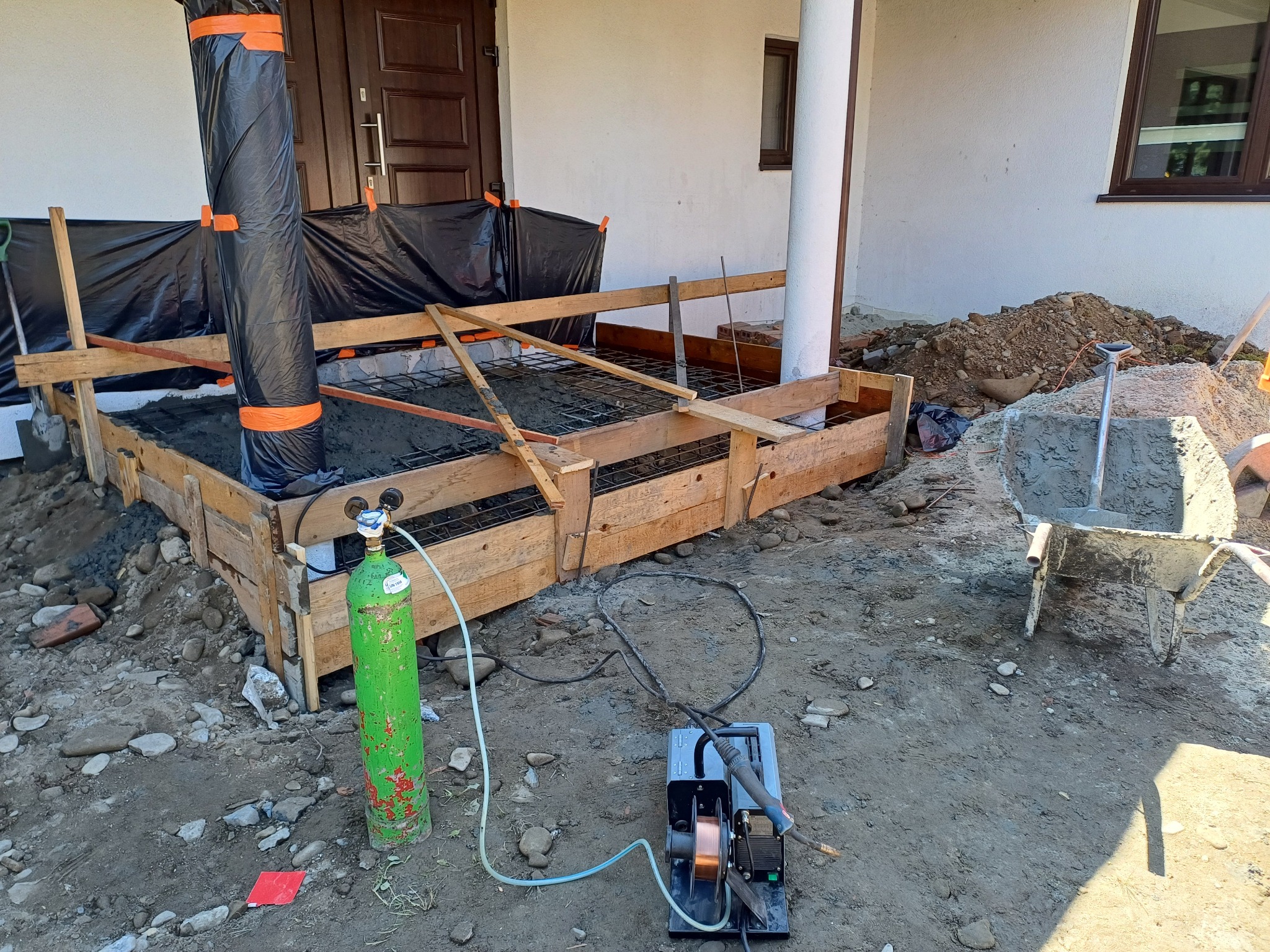 Przygotowanie fundamentów: drewniany szalunek, zbrojenie, częściowo zalany betonem. Butla z gazem i spawarka obok. Widok na wejście do domu i teren budowy.