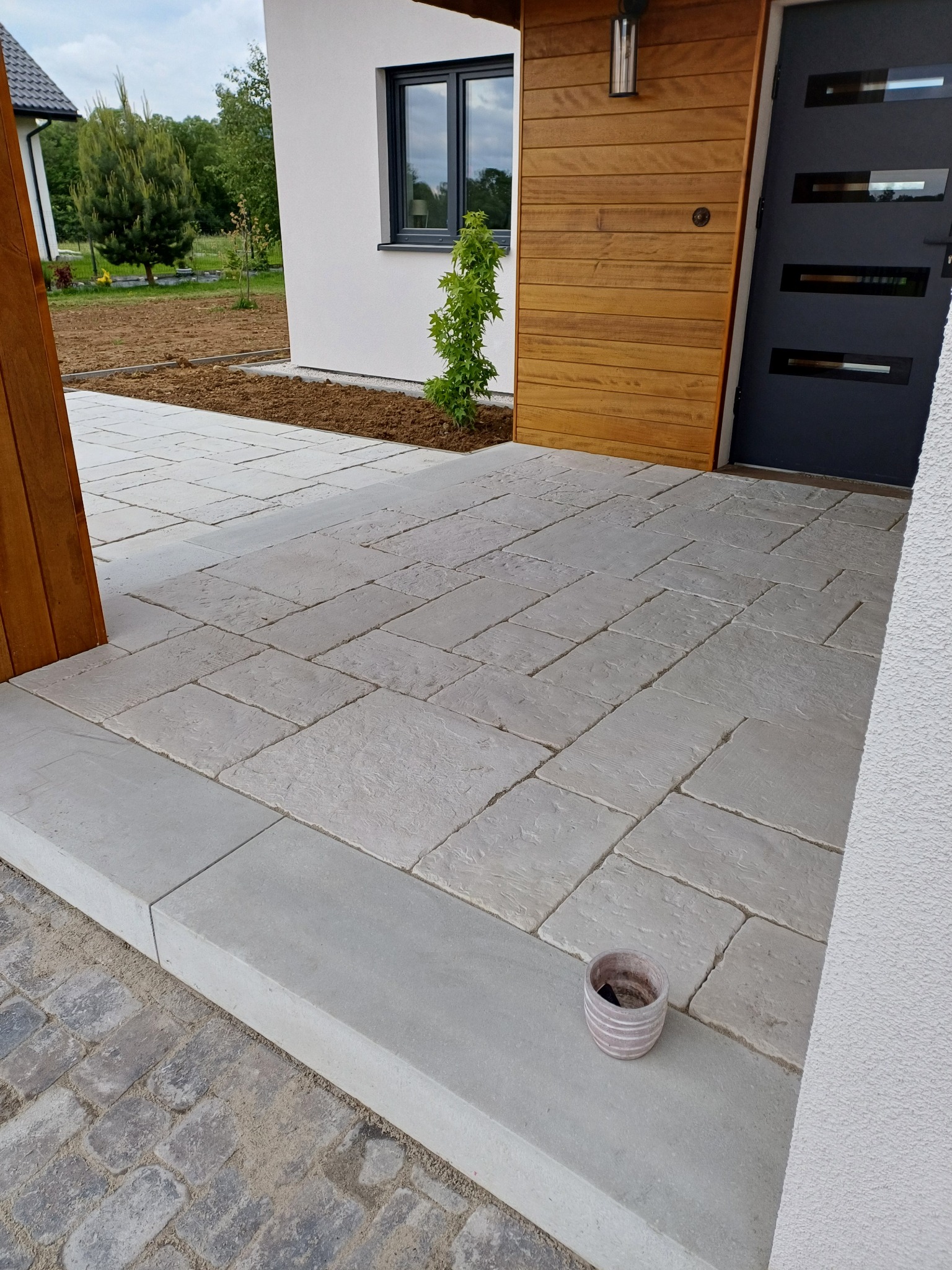 Jasnoszara kostka brukowa ułożona na podjeździe domu z drewnianą elewacją. Widoczny fragment rabaty i doniczka. Ujęcie z bliska, podkreślające wzór i fakturę kostki.