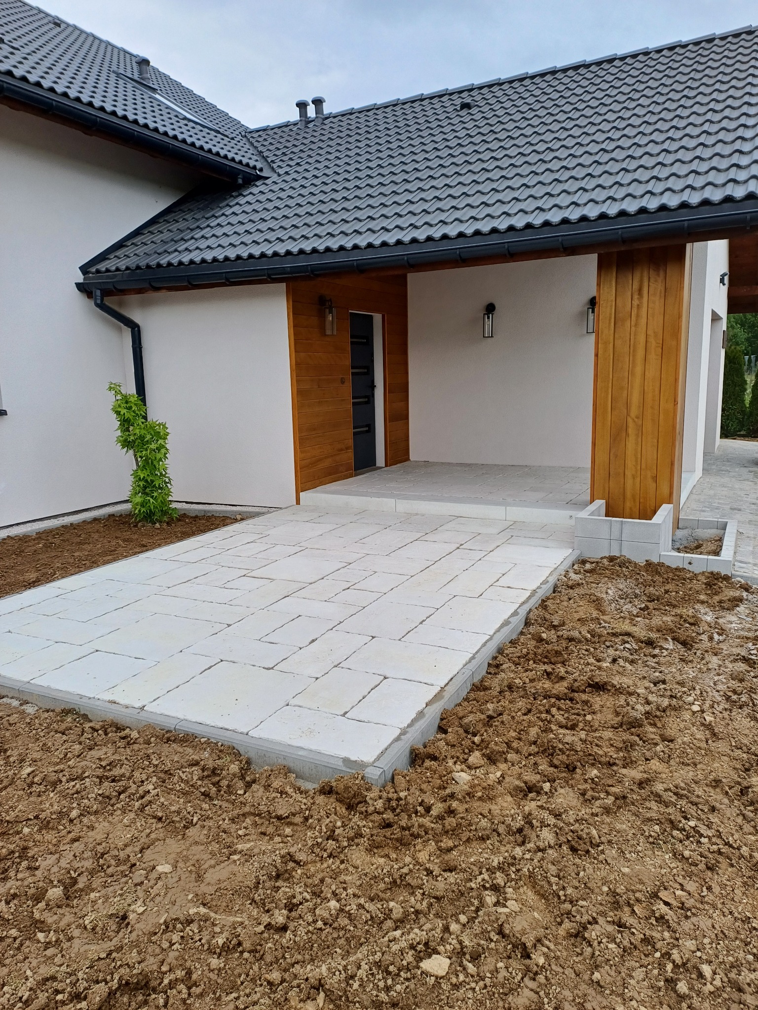 Nowoczesny podjazd z jasnej kostki brukowej przy domu z drewnianymi elementami. Widoczne prace ziemne i przygotowanie terenu pod dalsze prace brukarskie.