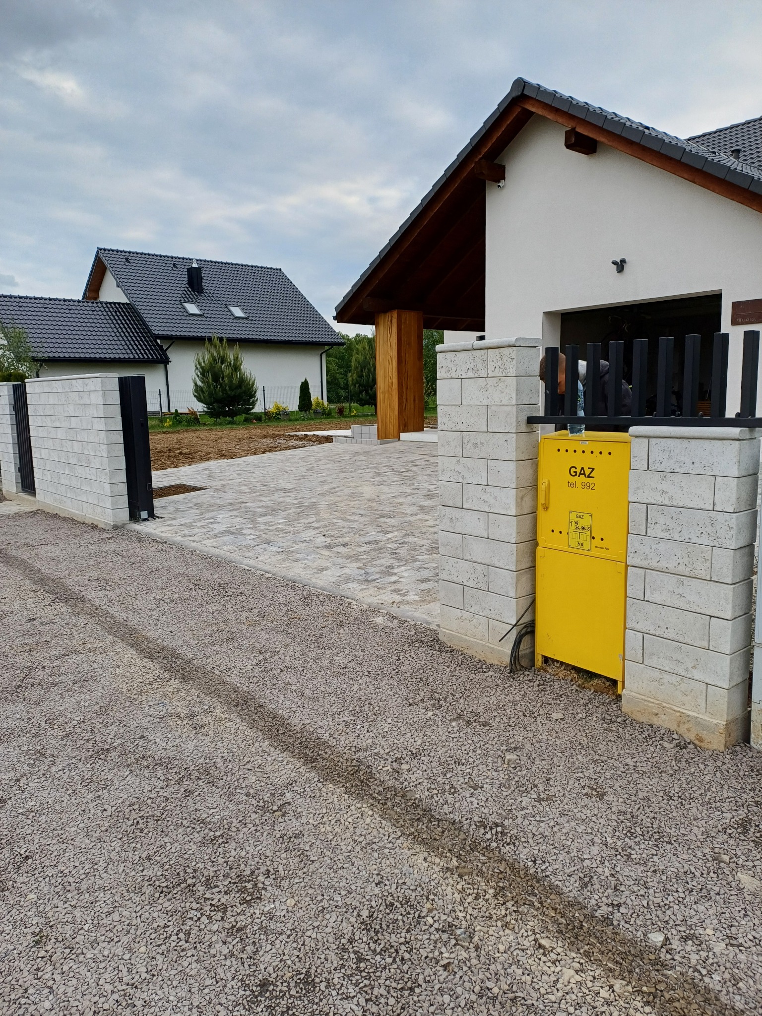 Nowoczesny podjazd z kostki brukowej prowadzący do domu z garażem, widoczna brama wjazdowa z elementami ogrodzenia i skrzynka gazowa.