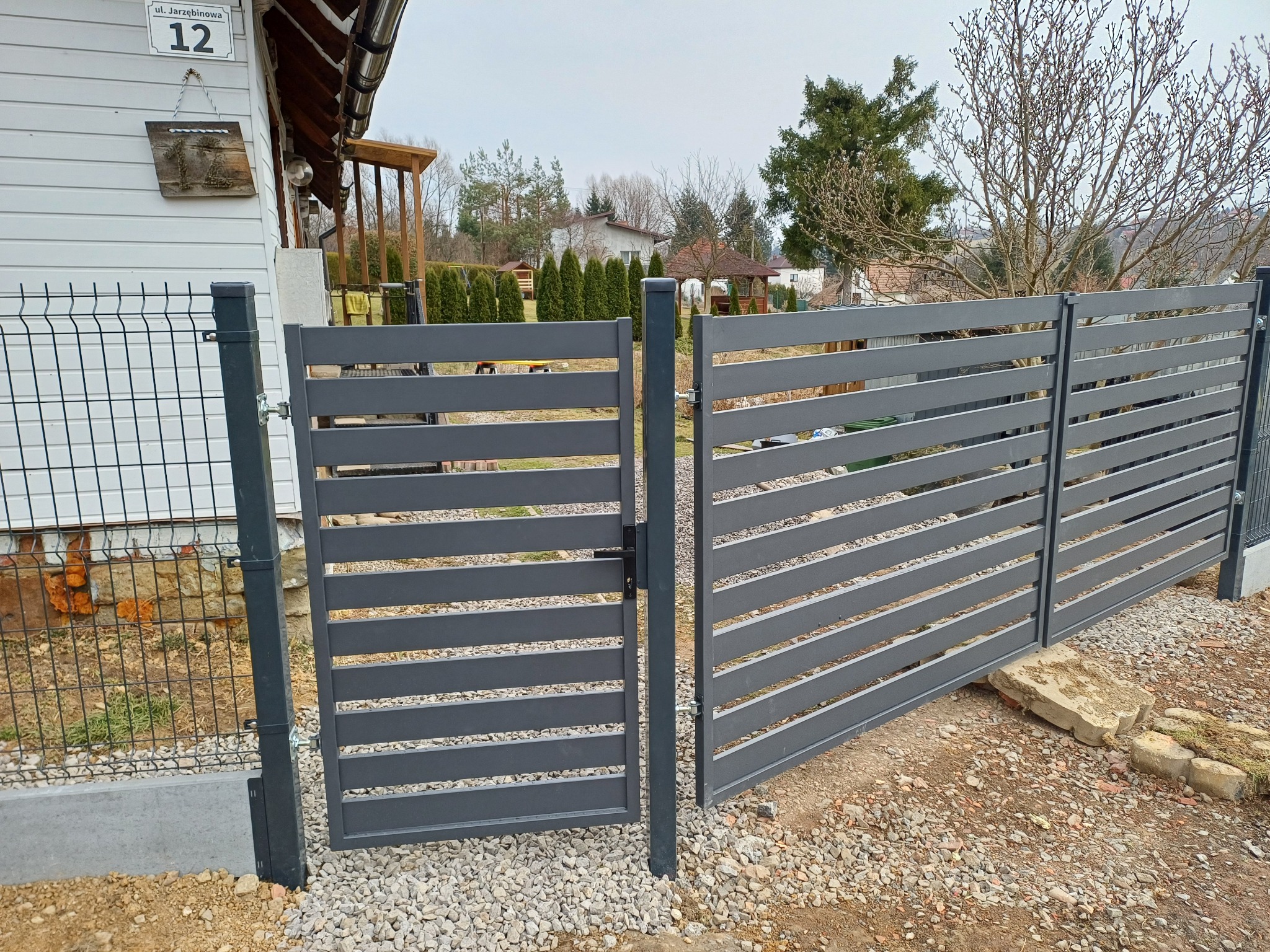 Nowoczesne, poziome ogrodzenie panelowe w kolorze grafitowym z furtką, zamontowane na posesji z widokiem na dom i ogród. Widoczny fragment siatki ogrodzeniowej.