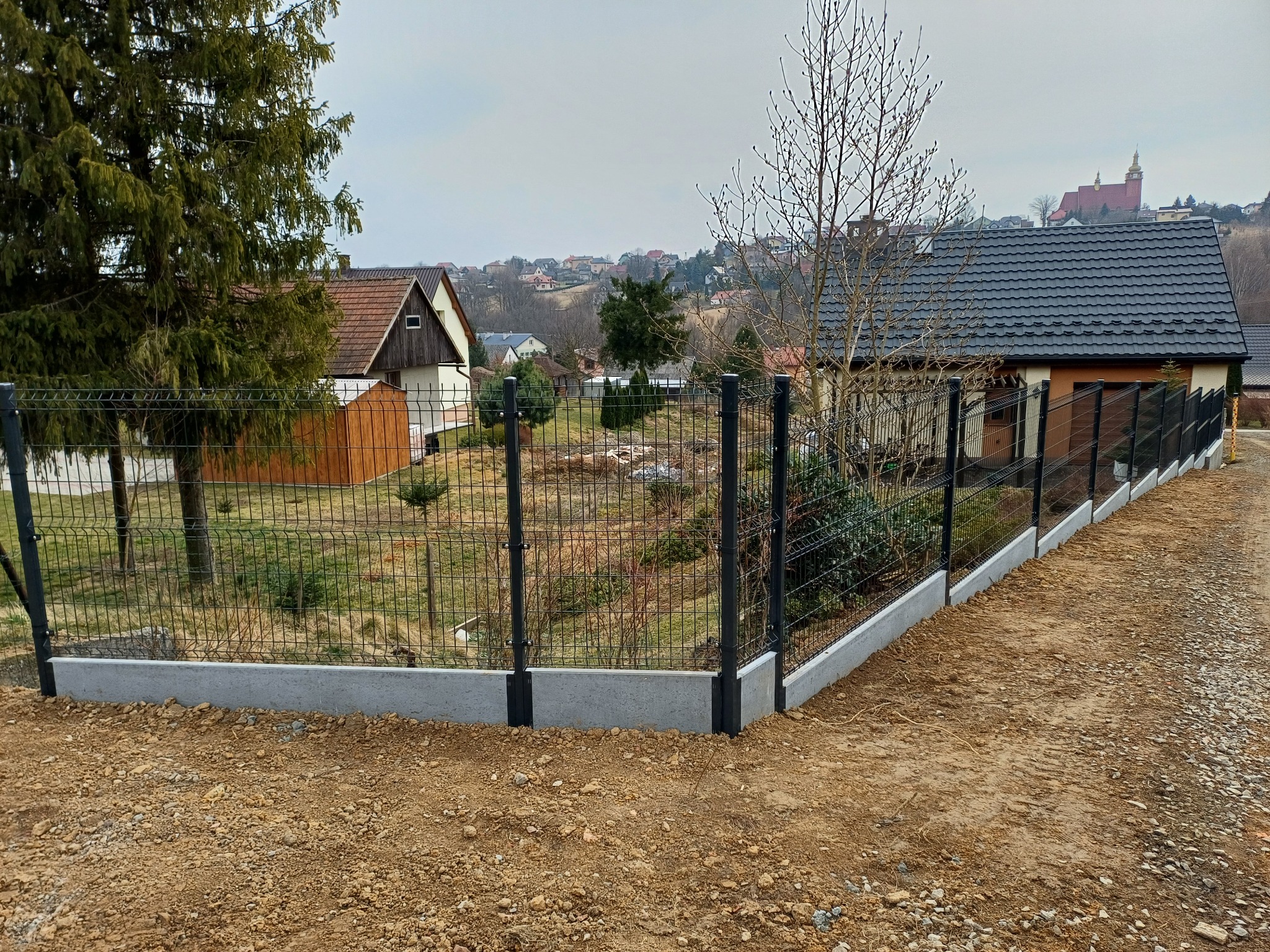 Nowoczesne ogrodzenie panelowe z podmurówką betonową na nierównym terenie. W tle domy i kościół na wzgórzu. Szara kolorystyka, pochmurny dzień. Widok z poziomu gruntu.