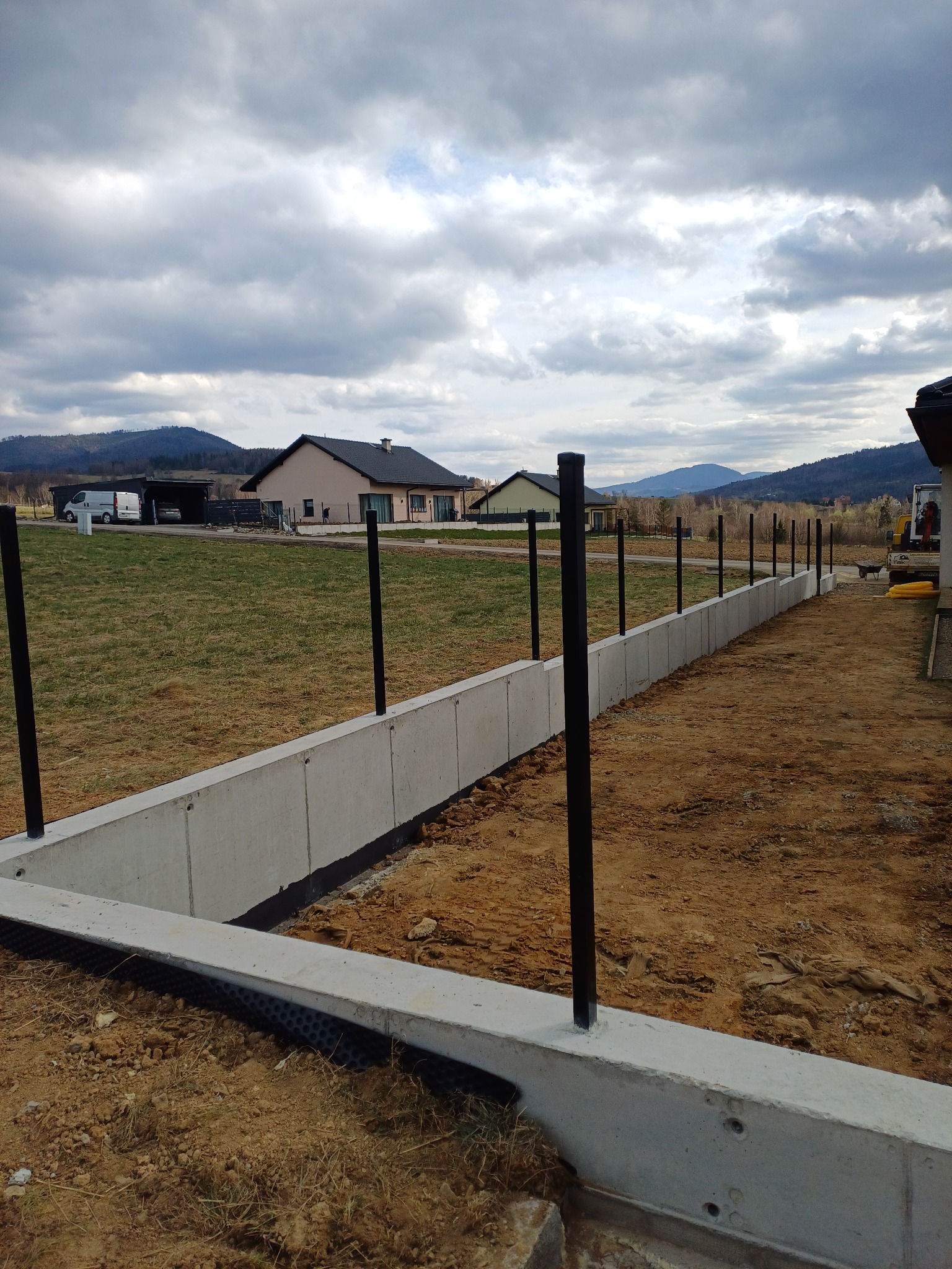 Betonowa podmurówka ogrodzenia z czarnymi słupkami wkopanymi w ziemię. W tle domy i góry pod pochmurnym niebem.