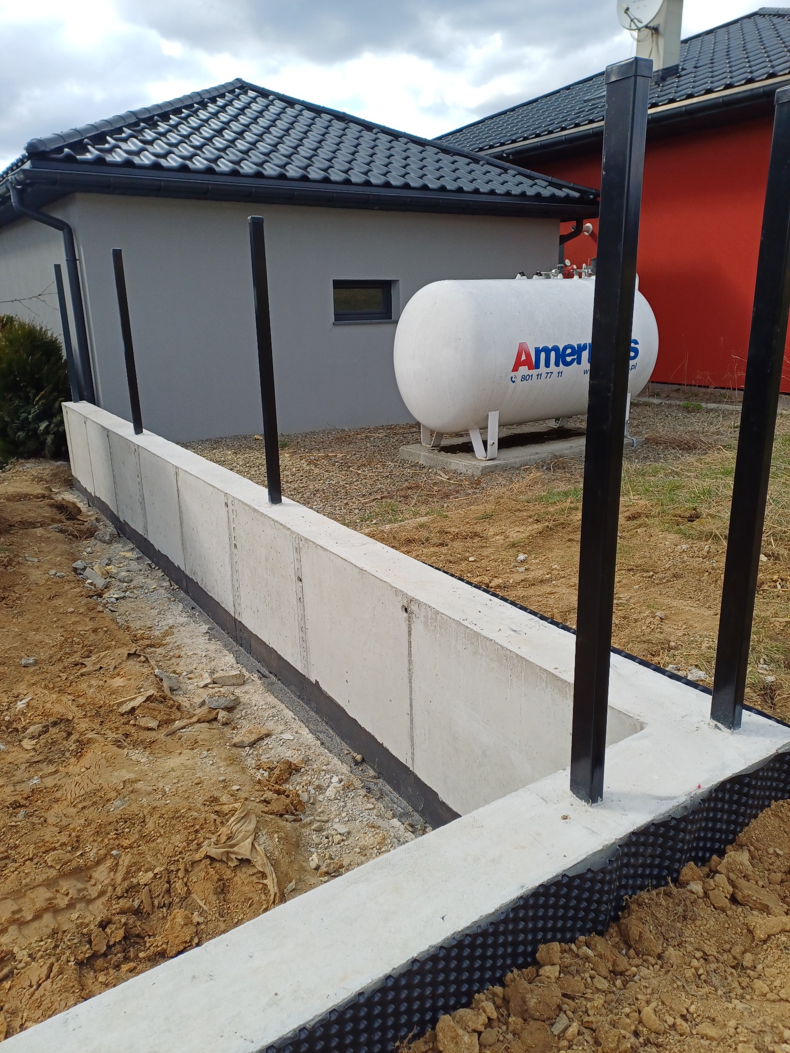 Betonowa podmurówka ogrodzenia z czarnymi słupkami na tle domu z zbiornikiem gazu. Widoczne zabezpieczenie hydroizolacyjne fundamentu.