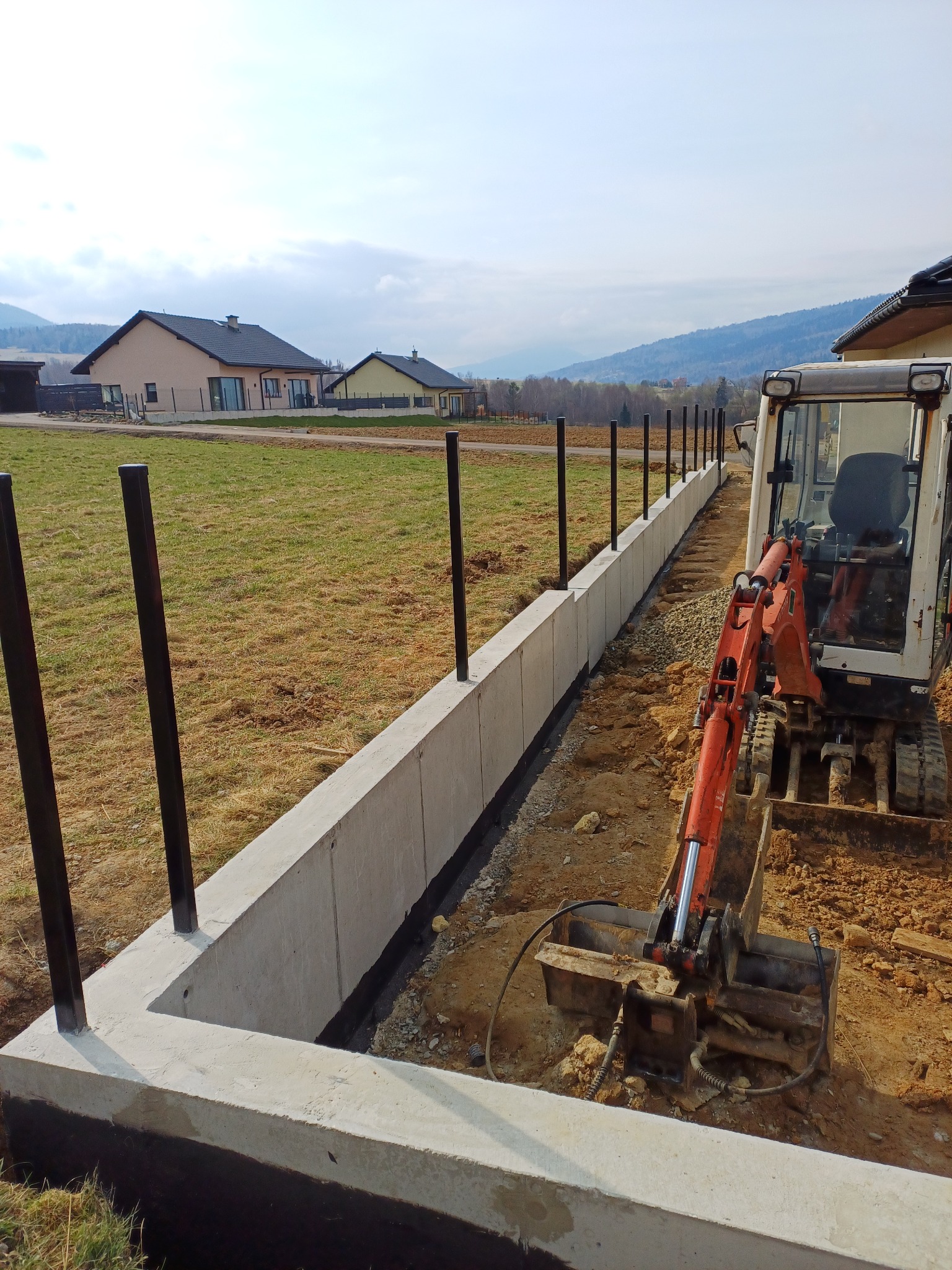 Fundament pod ogrodzenie z metalowymi słupkami w trakcie budowy, widoczna koparka, w tle domy i pagórkowaty krajobraz.