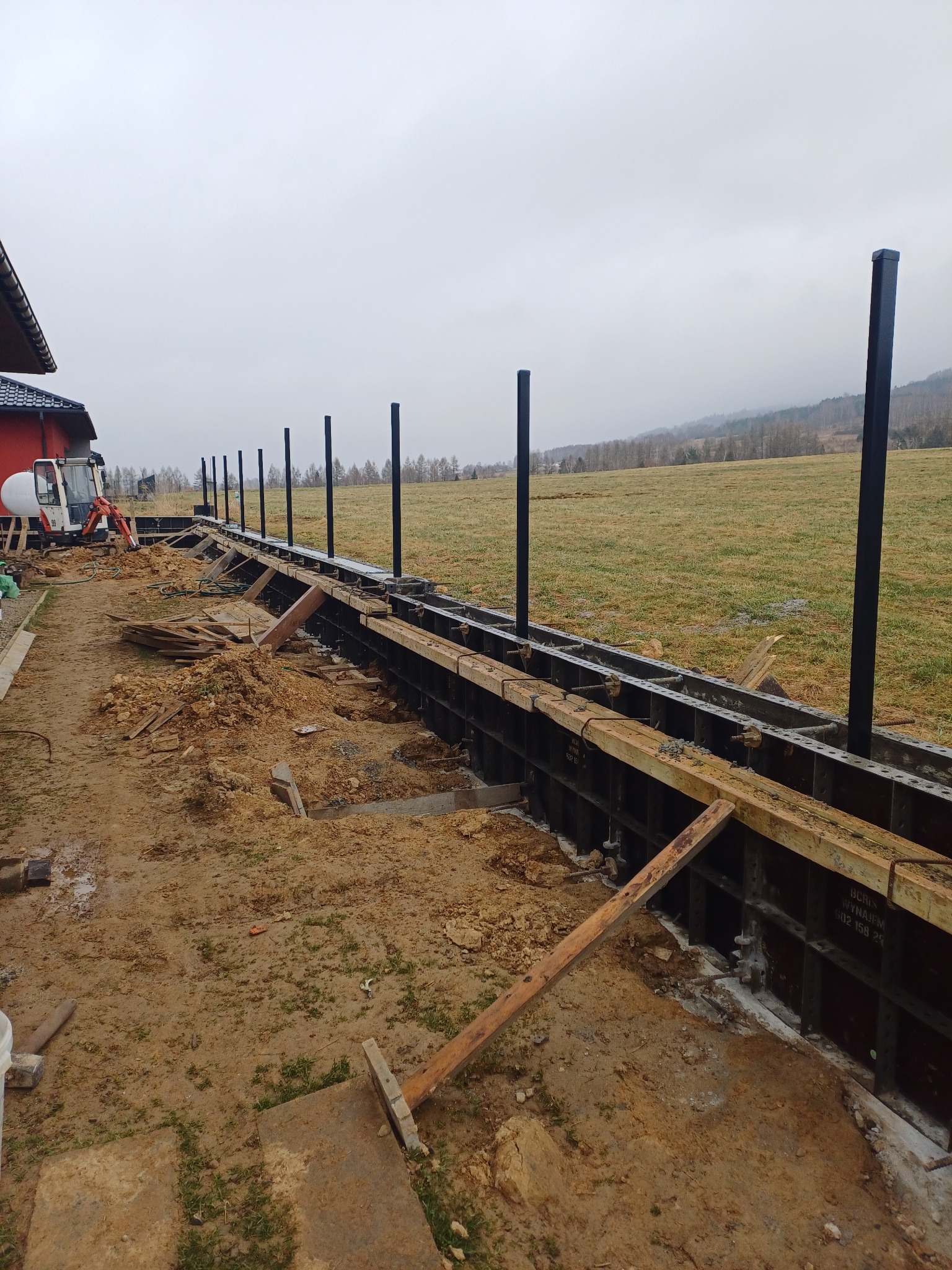 Budowa ogrodzenia: szalunek z metalowymi słupkami na fundamencie, w tle koparka i pagórkowaty krajobraz. Etap wylewania fundamentu pod ogrodzenie.