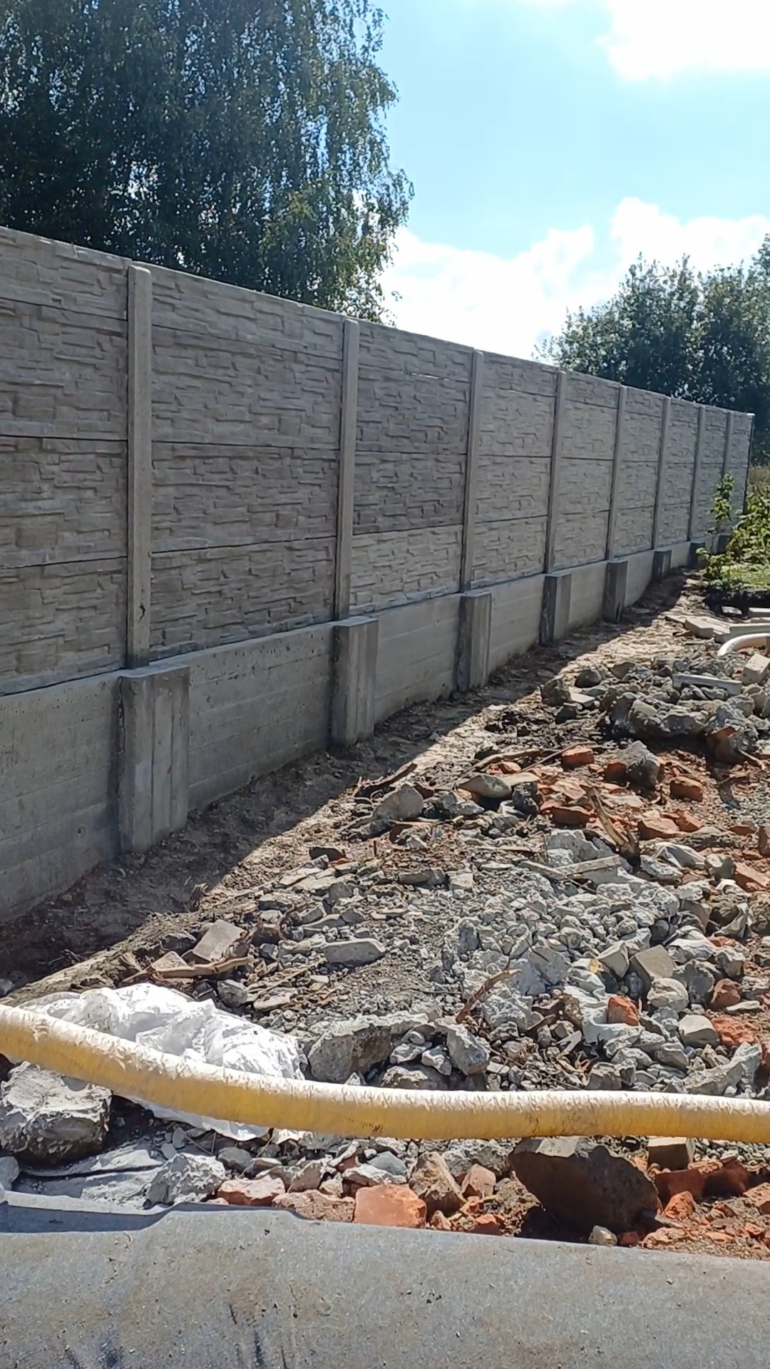 Długi, szary, prefabrykowany betonowy płot z fakturą kamienia, z widocznymi fundamentami, w trakcie montażu. Na pierwszym planie gruz i resztki materiałów budowlanych.