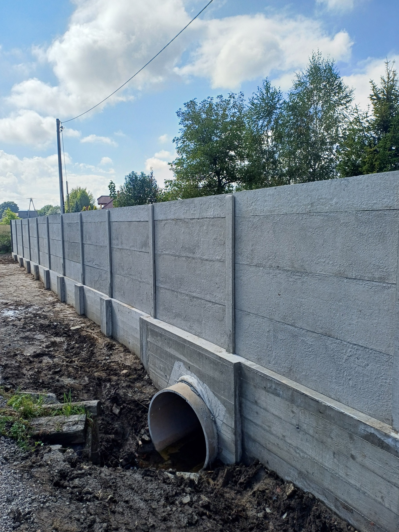 Długi, betonowy płot panelowy z rurą drenażową w ziemi, widok z boku. Tło: niebo z chmurami i drzewa. Elementy są szare, ziemia brązowa, a niebo błękitne.