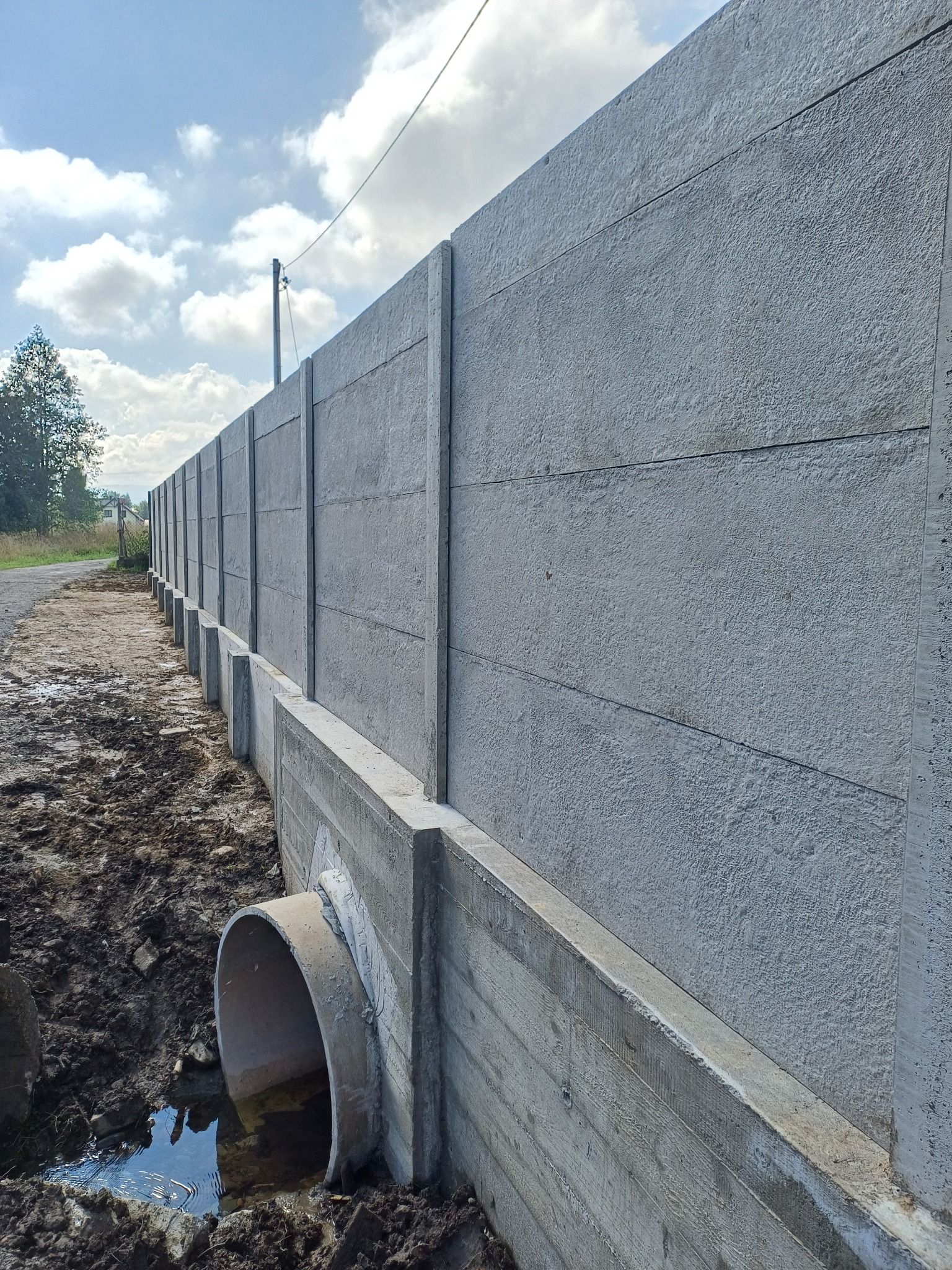 Długi, szary, betonowy mur oporowy z rurą drenażową, biegnący wzdłuż drogi, z widocznym niebem i drzewami w tle.