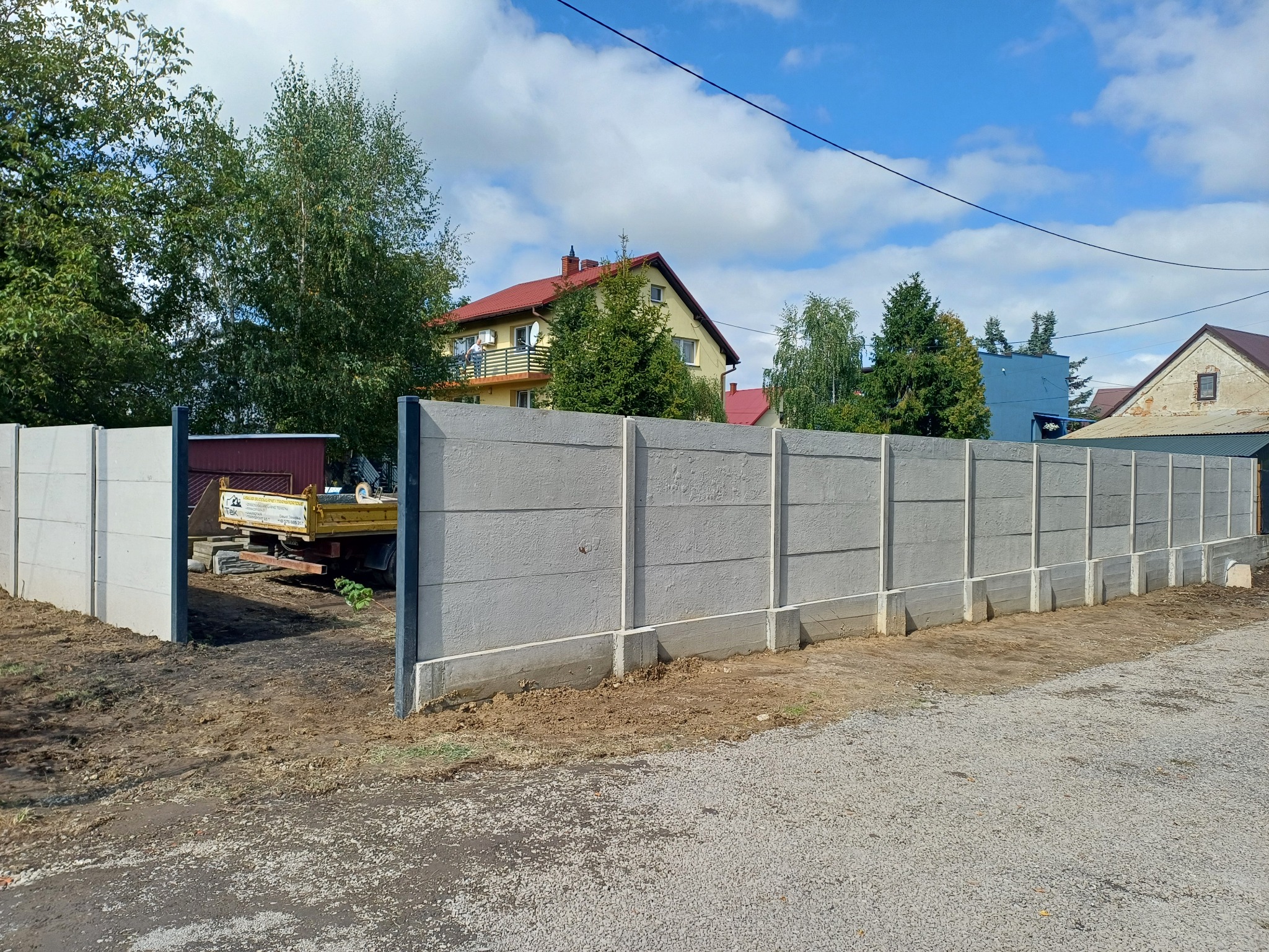 Nowo postawiony, betonowy płot panelowy w szarej tonacji, z widocznym samochodem dostawczym w tle i fragmentem domu z czerwonym dachem.