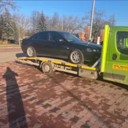 Czarny samochód osobowy na żółtej lawecie z zieloną kabiną pomocy drogowej, widok z boku, na pierwszym planie cień osoby fotografującej na kostce brukowej.