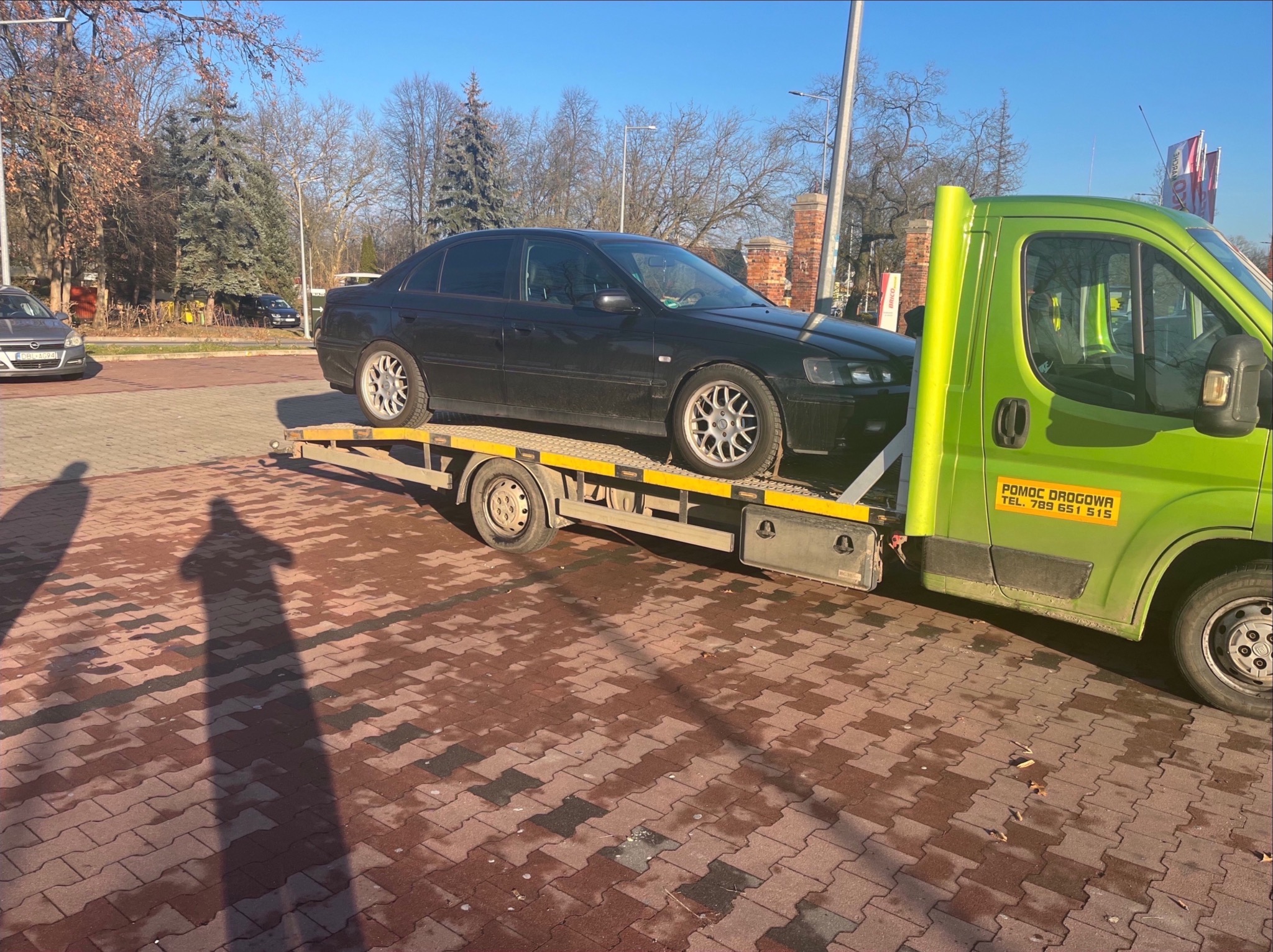 Czarny samochód osobowy na żółtej lawecie z zieloną kabiną pomocy drogowej, widok z boku, na pierwszym planie cień osoby fotografującej na kostce brukowej.