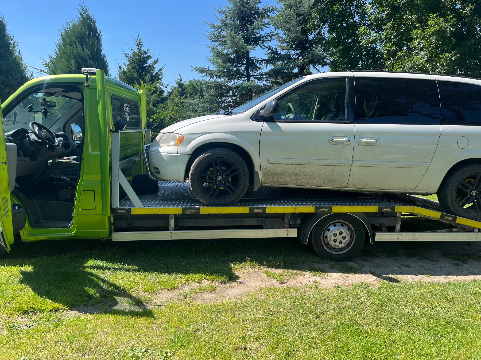 Jasnozielona ciężarówka z otwartymi drzwiami kabiny przewozi na platformie biały samochód osobowy typu minivan na tle zielonych drzew.