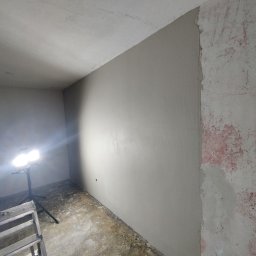 P.R - Ściana po nałożeniu gładzi szpachlowej, z widocznymi śladami wcześniejszego malowania na sąsiedniej ścianie, oświetlona lampą roboczą na statywie, drabina malarska w rogu.