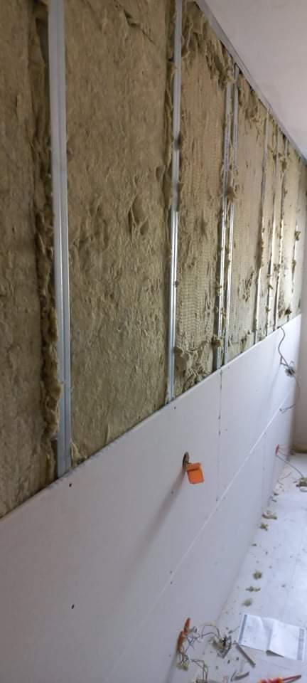 Ściana w trakcie wygłuszania wełną mineralną, z zamontowanymi metalowymi profilami i częściowo przytwierdzonymi płytami gipsowo-kartonowymi, widoczny otwór na puszkę elektryczną.
