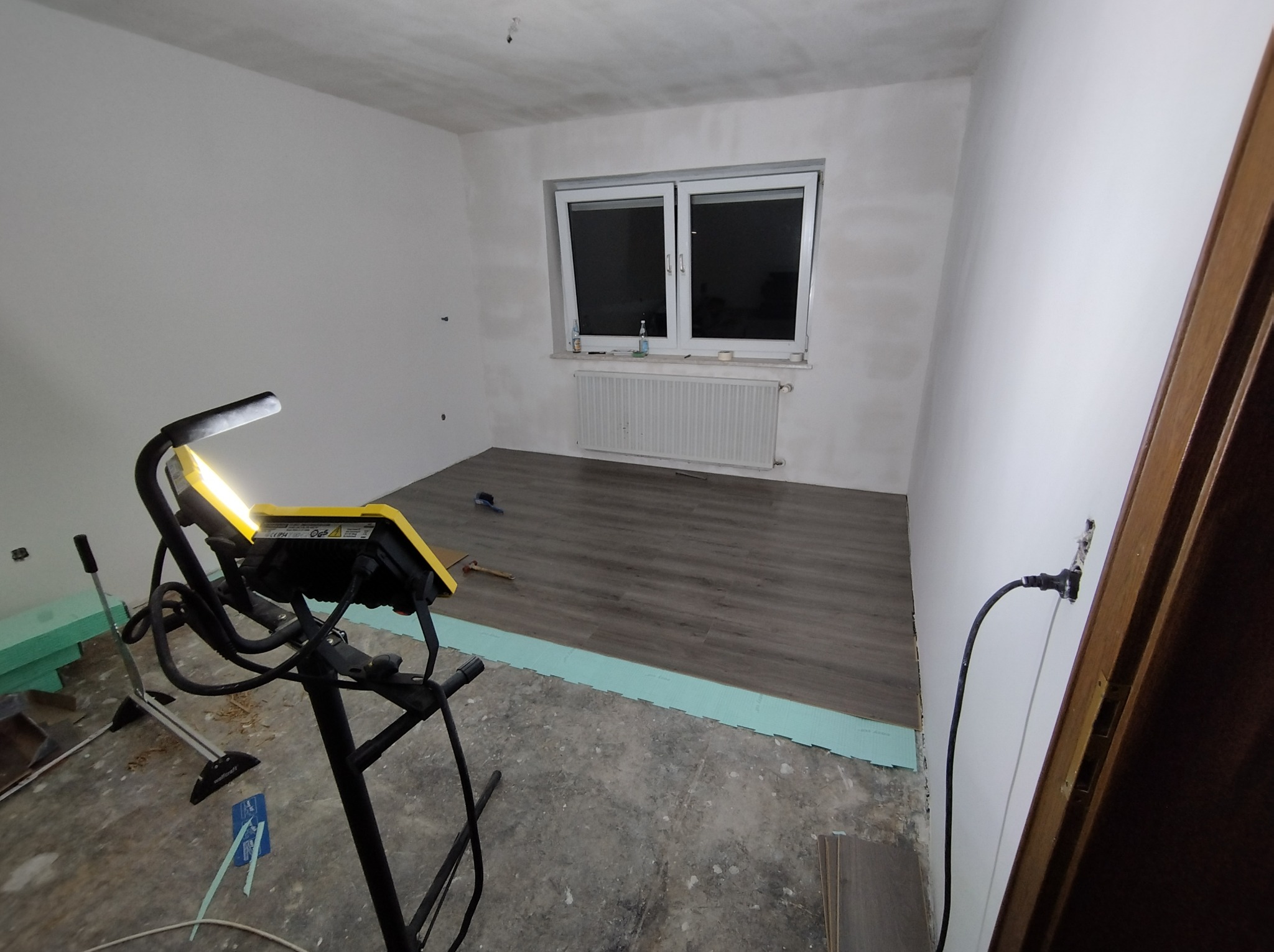 Montaż paneli podłogowych w pokoju: widoczny fragment ułożonej podłogi, zielona pianka izolacyjna, lampa budowlana na statywie, surowe ściany przygotowane do malowania, okno z grzejnikiem.