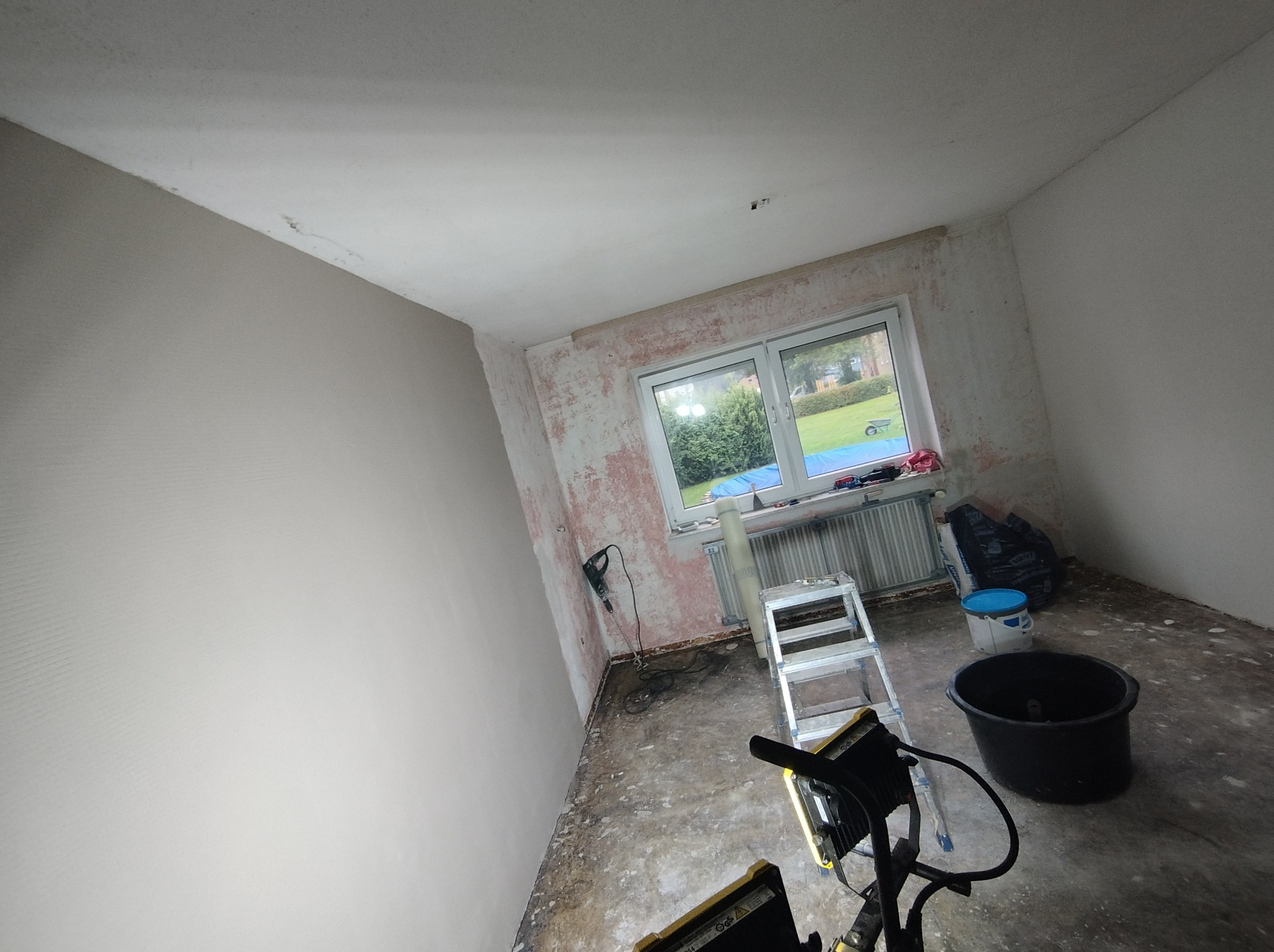 Pokój w trakcie prac przygotowawczych do malowania: ściany częściowo zagruntowane, widoczne ślady po usuniętej farbie, drabina, wiadro, szpachla elektryczna i inne narzędzia malarskie.
