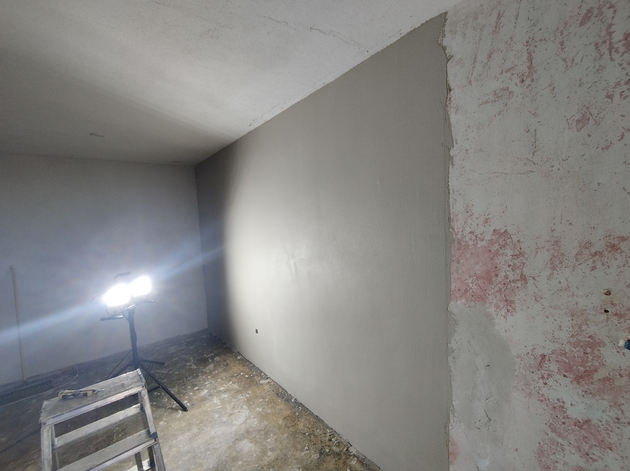 Ściana po nałożeniu gładzi szpachlowej, z widocznymi śladami wcześniejszego malowania na sąsiedniej ścianie, oświetlona lampą roboczą na statywie, drabina malarska w rogu.