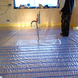 D.S.ELEKTRO DAMIAN STRUMIŁŁO - Rozłożona instalacja ogrzewania podłogowego z niebieskimi rurkami na folii, widoczne narzędzia i fragment osoby w pomieszczeniu z drewnianymi ścianami.