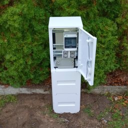 D.S.ELEKTRO DAMIAN STRUMIŁŁO - Biała skrzynka elektryczna z otwartymi drzwiczkami, umieszczona na zewnątrz, na tle zielonych krzewów. Widoczne elementy instalacji wewnątrz.