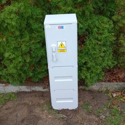 D.S.ELEKTRO DAMIAN STRUMIŁŁO - Szara skrzynka elektryczna z ostrzeżeniem o wysokim napięciu, umieszczona na ziemi przed zielonym żywopłotem. Widoczne narzędzia i opakowania po montażu.