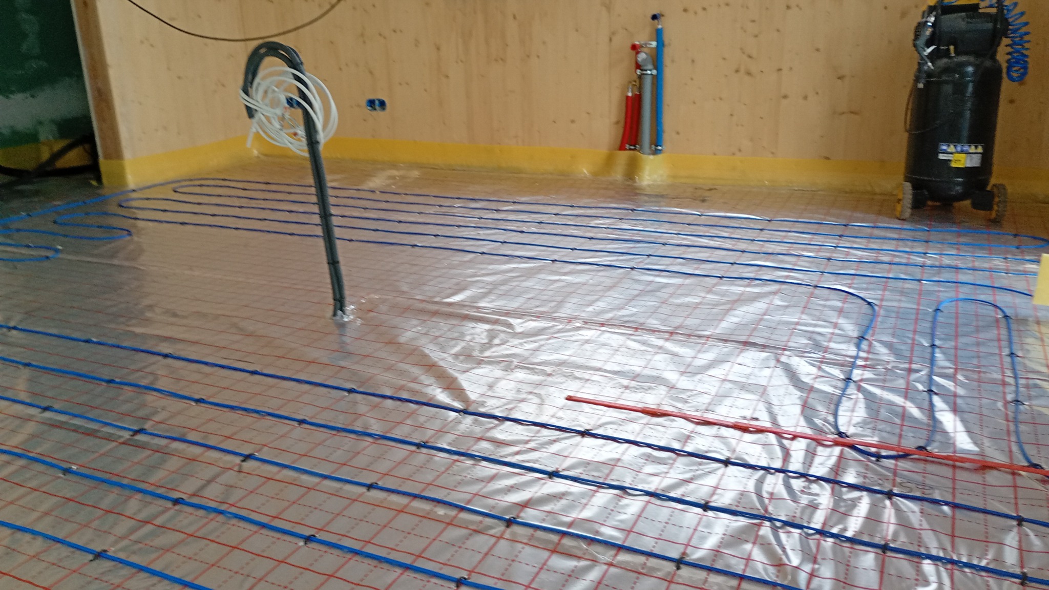 Ułożone rury ogrzewania podłogowego na folii aluminiowej w pomieszczeniu z drewnianymi ścianami. Widoczny kompresor i pionowe rury przy ścianie.