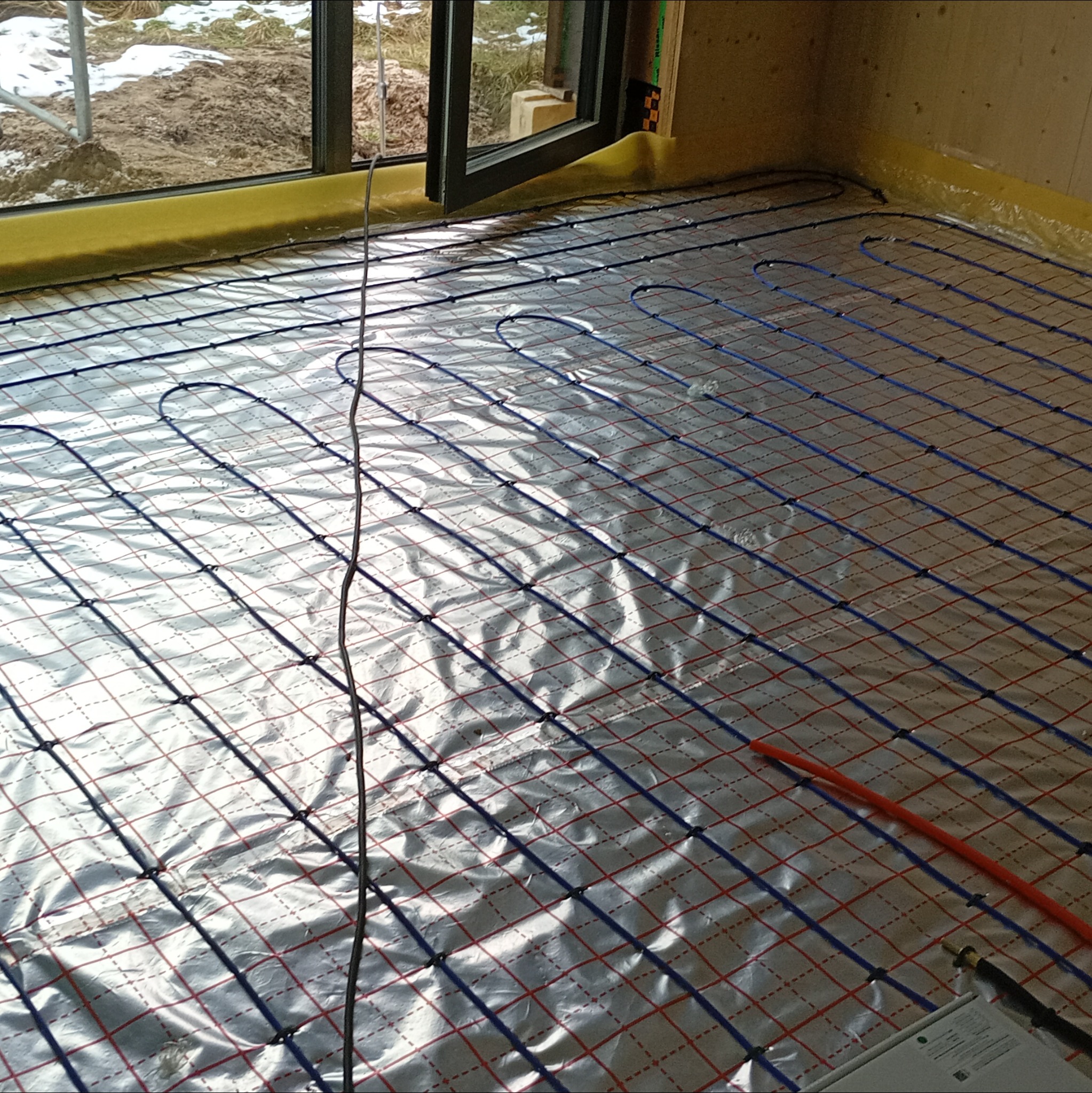 Ułożona instalacja ogrzewania podłogowego na folii aluminiowej z widocznymi niebieskimi rurkami, zabezpieczona żółtą folią przy ścianie. Widok z góry.