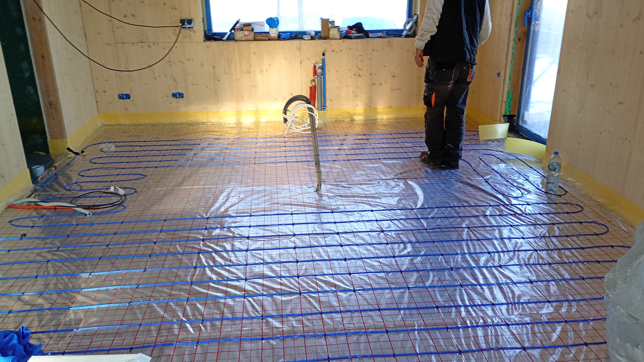 Rozłożona instalacja ogrzewania podłogowego z niebieskimi rurkami na folii, widoczne narzędzia i fragment osoby w pomieszczeniu z drewnianymi ścianami.