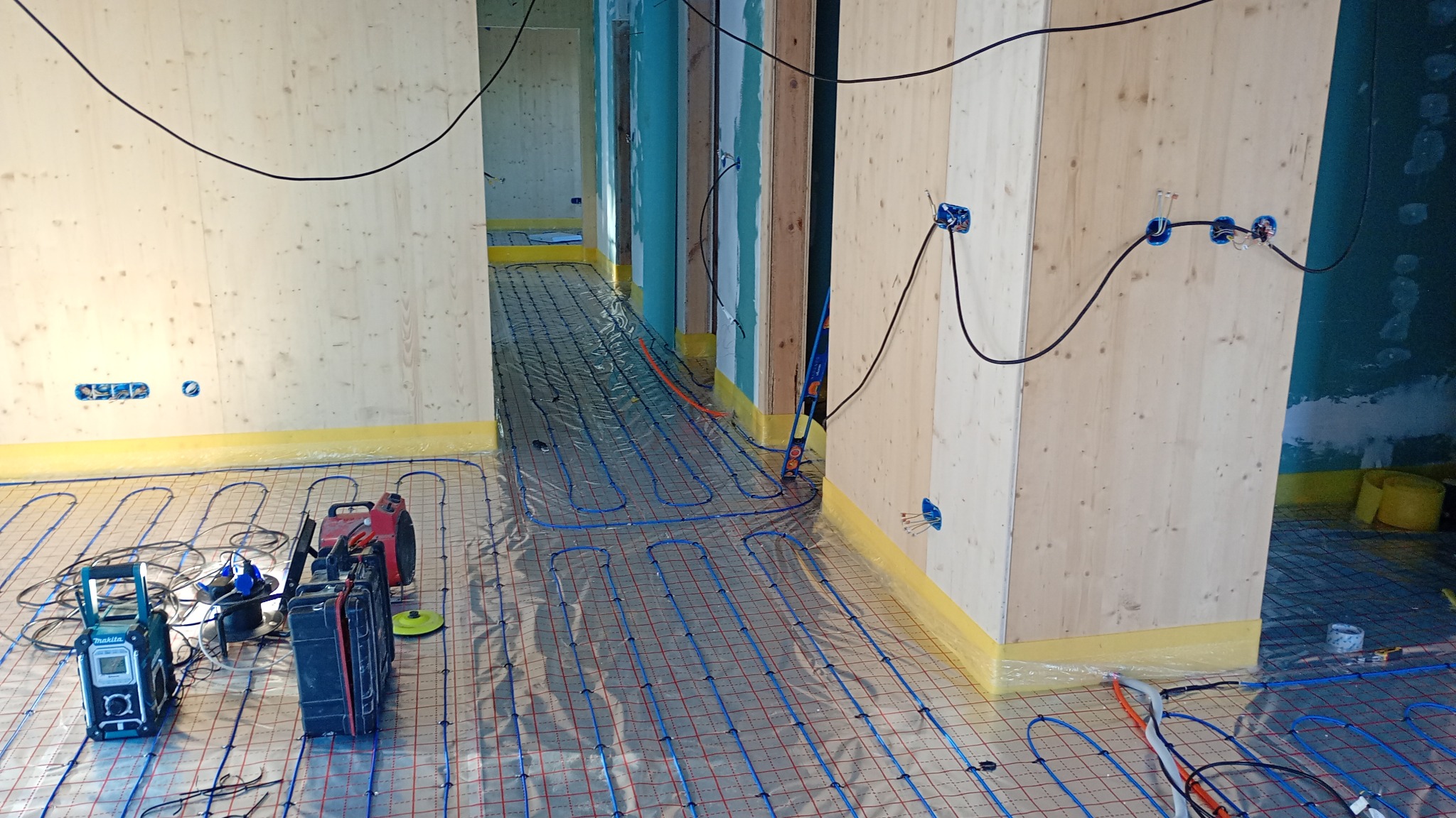 Montaż ogrzewania podłogowego: rury grzewcze ułożone na folii, widoczne narzędzia i kable elektryczne w surowym wnętrzu z drewnianymi ścianami i instalacją elektryczną.