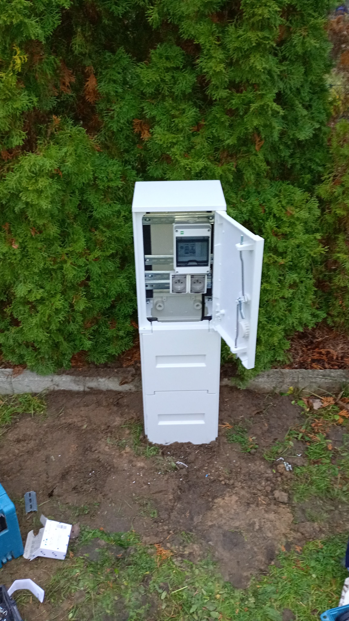 Biała skrzynka elektryczna z otwartymi drzwiczkami, umieszczona na zewnątrz, na tle zielonych krzewów. Widoczne elementy instalacji wewnątrz.