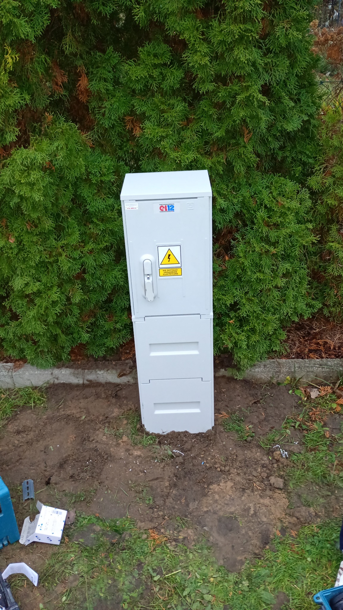 Szara skrzynka elektryczna z ostrzeżeniem o wysokim napięciu, umieszczona na ziemi przed zielonym żywopłotem. Widoczne narzędzia i opakowania po montażu.