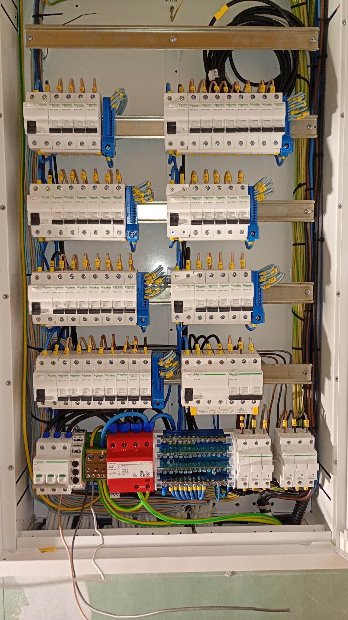 Rozdzielnica elektryczna Schneider z zabezpieczeniami, widoczne liczne przewody w kolorach: niebieskim, żółtym, zielonym i brązowym. Elementy zamontowane w białej obudowie.