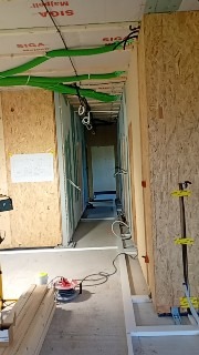 Wnętrze w trakcie budowy: widoczne ściany z płyt OSB, zielone rury wentylacyjne pod sufitem, przewody elektryczne i narzędzia na podłodze. Etap prac instalacyjnych.