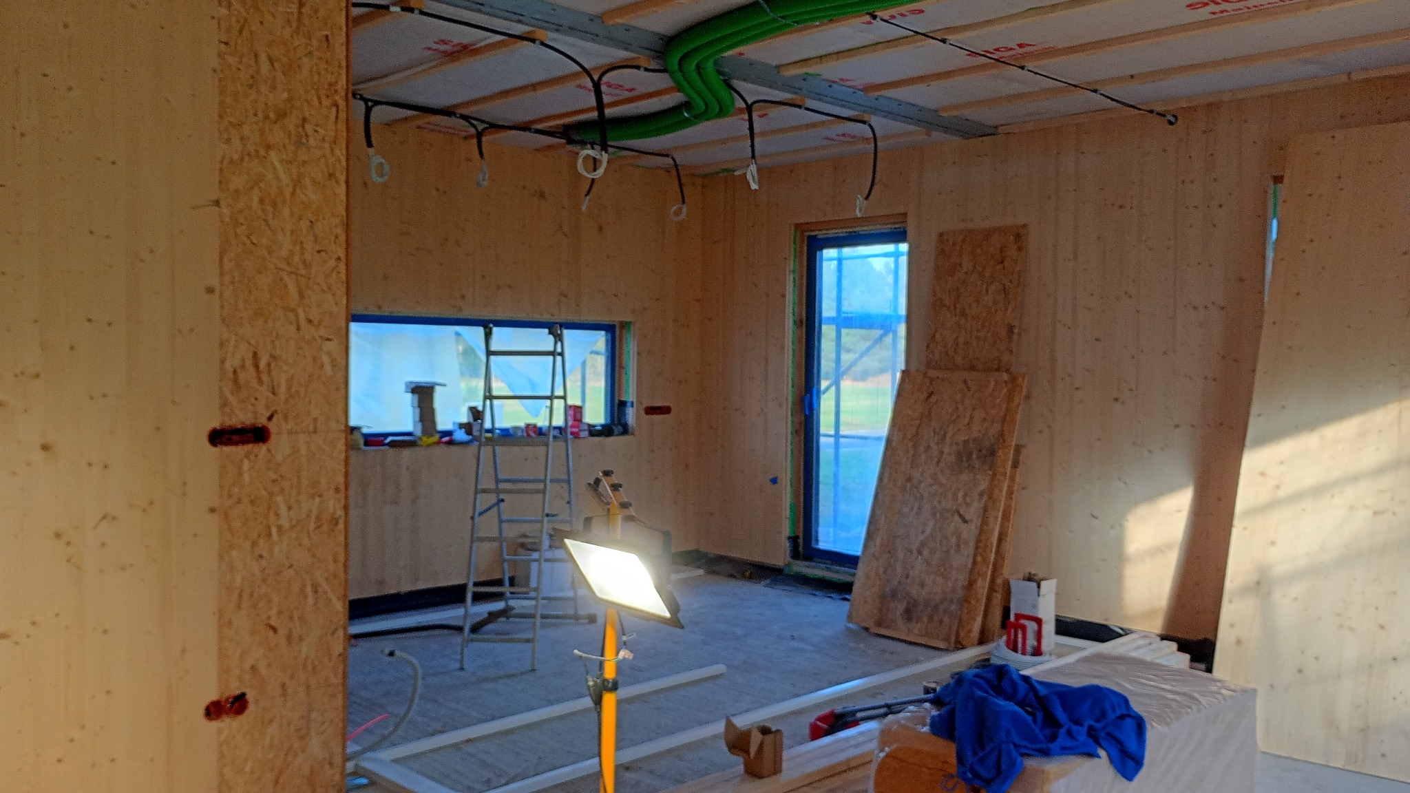 Wnętrze domu w trakcie budowy z widoczną instalacją elektryczną pod sufitem, oświetleniem roboczym i materiałami budowlanymi. Ściany wykończone drewnem.