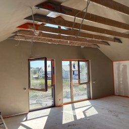 MikroEfekt Wiktor Herman - Wykończone wnętrze poddasza ze skosami, ściany i posadzka w kolorze NCS 2010-y30r, okna z niebieską ramą, widoczne drewniane belki stropowe.