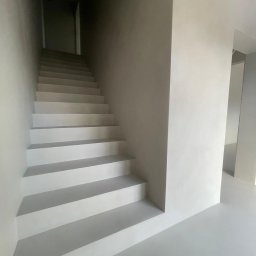 MikroEfekt Wiktor Herman - Minimalistyczne schody wewnętrzne w jasnej tonacji szarości, wykończone jednolitą powierzchnią, z subtelnym oświetleniem podkreślającym gładką strukturę.