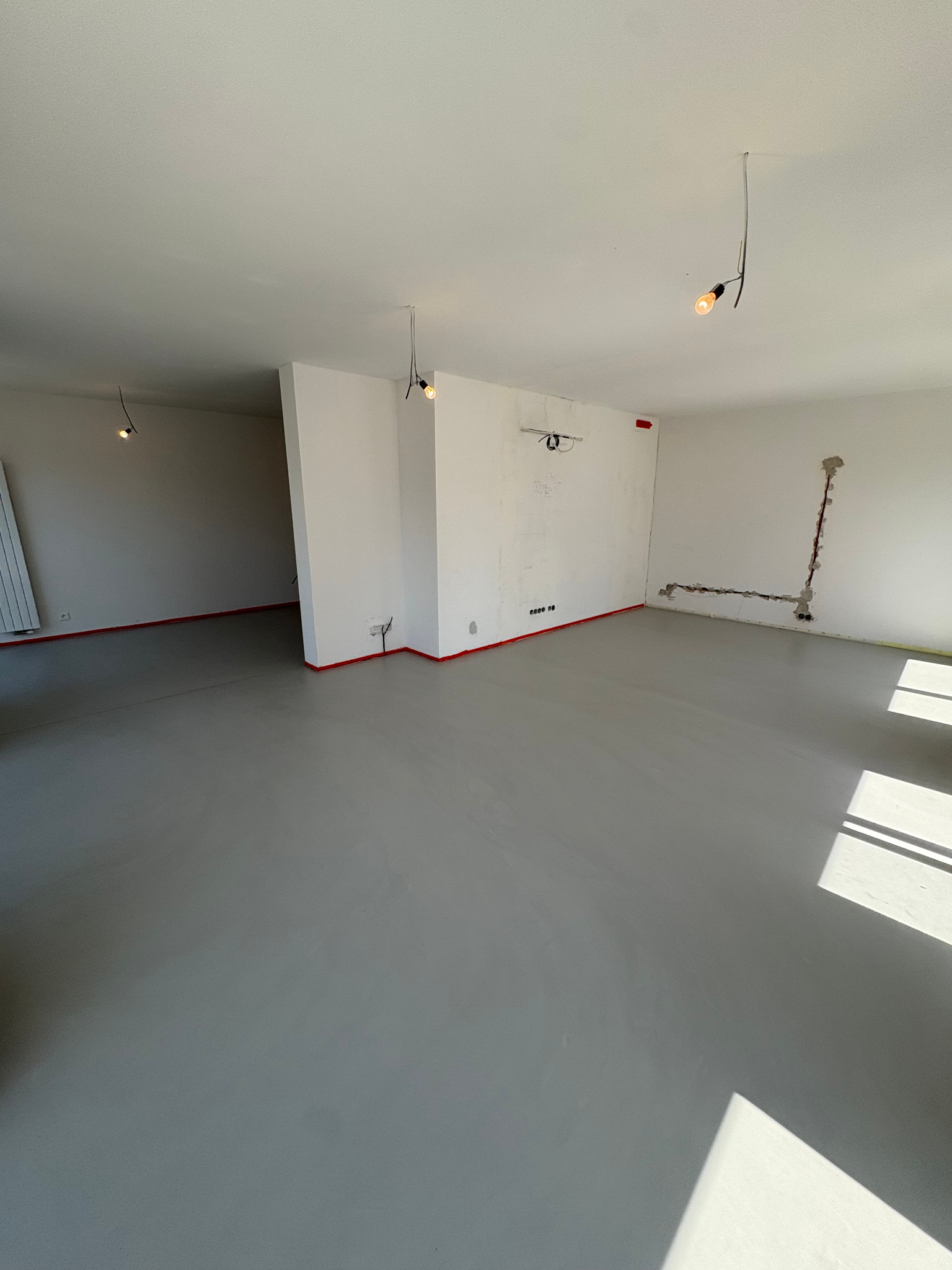 Szara posadzka w jasnym, pustym pomieszczeniu z widocznymi instalacjami elektrycznymi i oświetleniem punktowym. Czerwona taśma zabezpiecza krawędzie ścian.