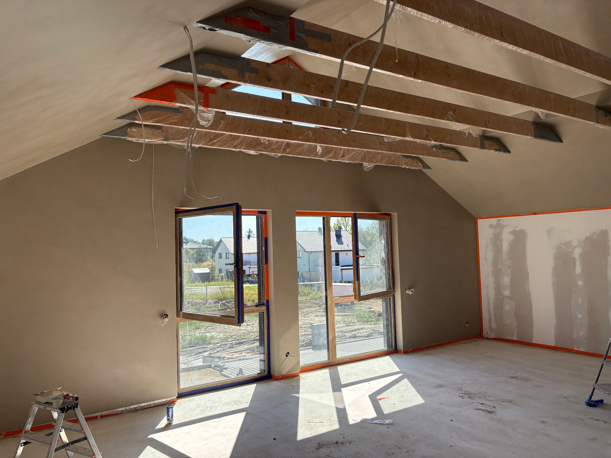 Wykończone wnętrze poddasza ze skosami, ściany i posadzka w kolorze NCS 2010-y30r, okna z niebieską ramą, widoczne drewniane belki stropowe.