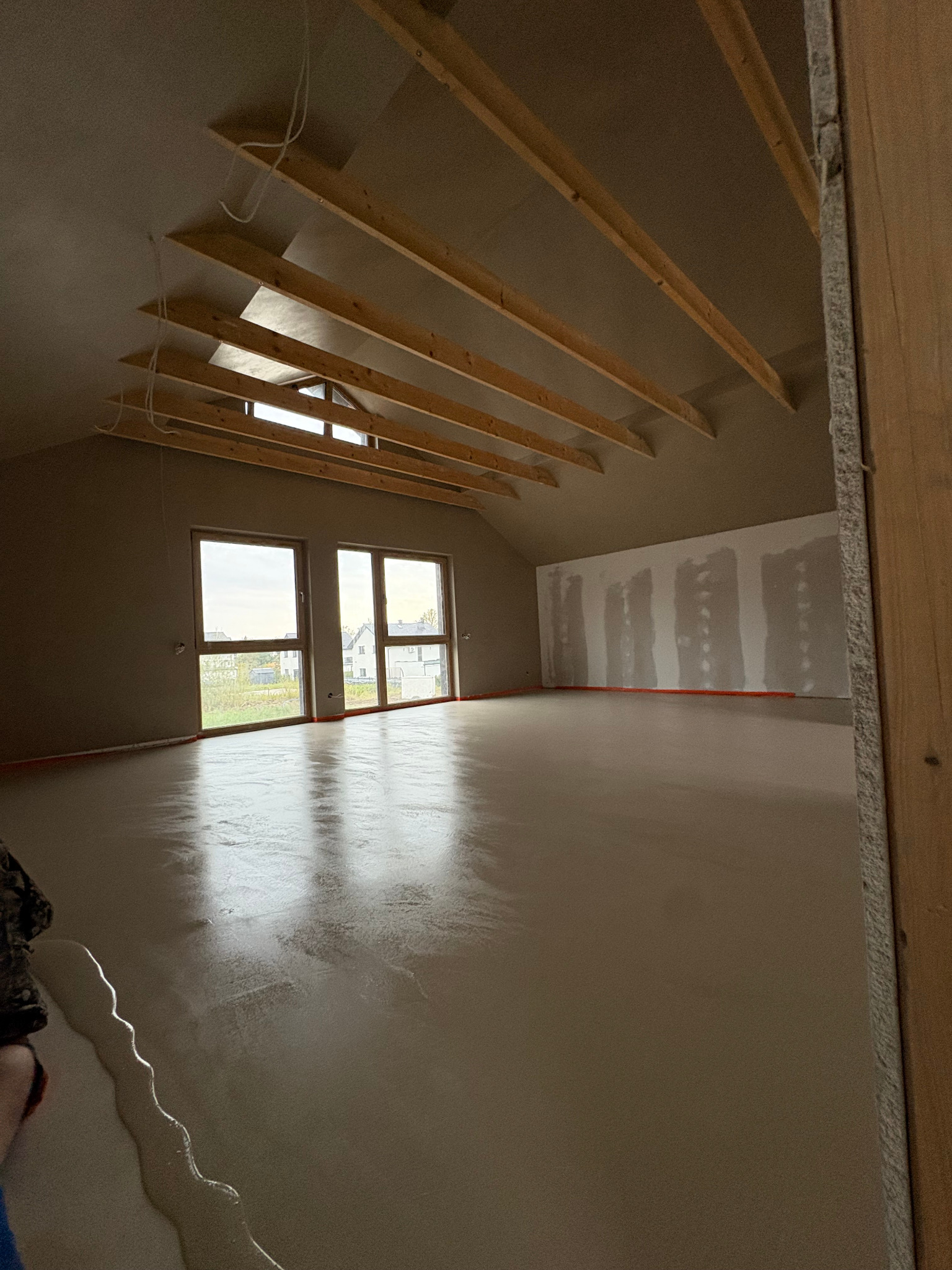 Nowa, jasna posadzka w odcieniach beżu w pomieszczeniu ze skosami i oknami. Widoczne drewniane elementy konstrukcyjne sufitu i fragment ściany z płyt gipsowych.