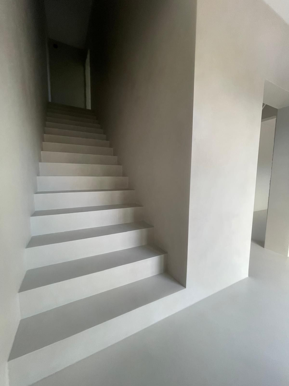 Minimalistyczne schody wewnętrzne w jasnej tonacji szarości, wykończone jednolitą powierzchnią, z subtelnym oświetleniem podkreślającym gładką strukturę.