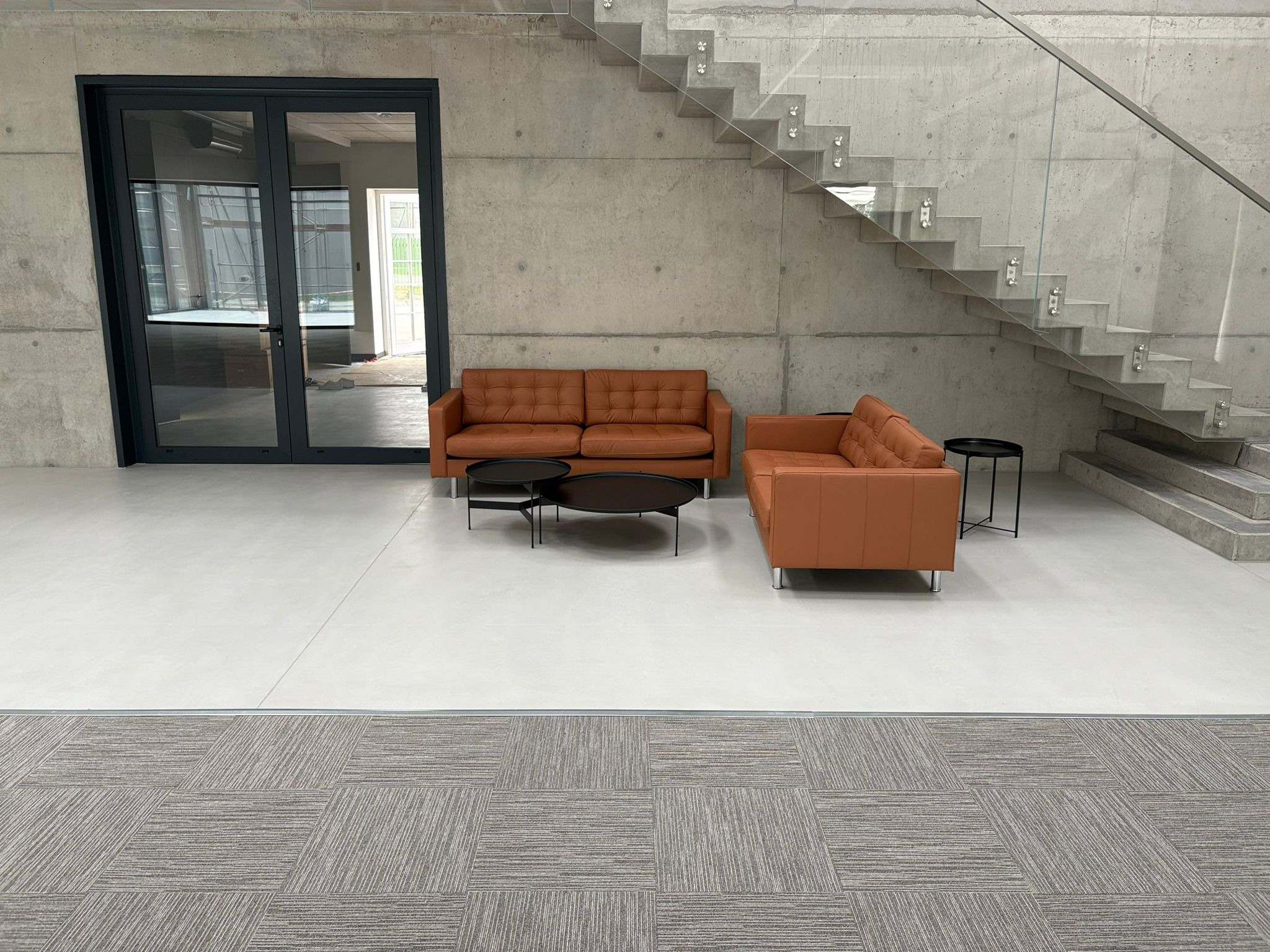 Jasna posadzka w systemie Quarelli hardpox w przestronnym wnętrzu z betonowymi ścianami, schodami i brązowymi sofami. Widok na wykończone podłogi w biurze.