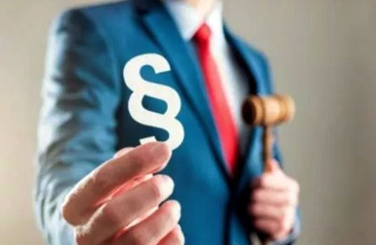 Mężczyzna w niebieskim garniturze i czerwonym krawacie trzyma w dłoni biały symbol paragrafu, a w drugiej ręce drewniany młotek sędziowski.